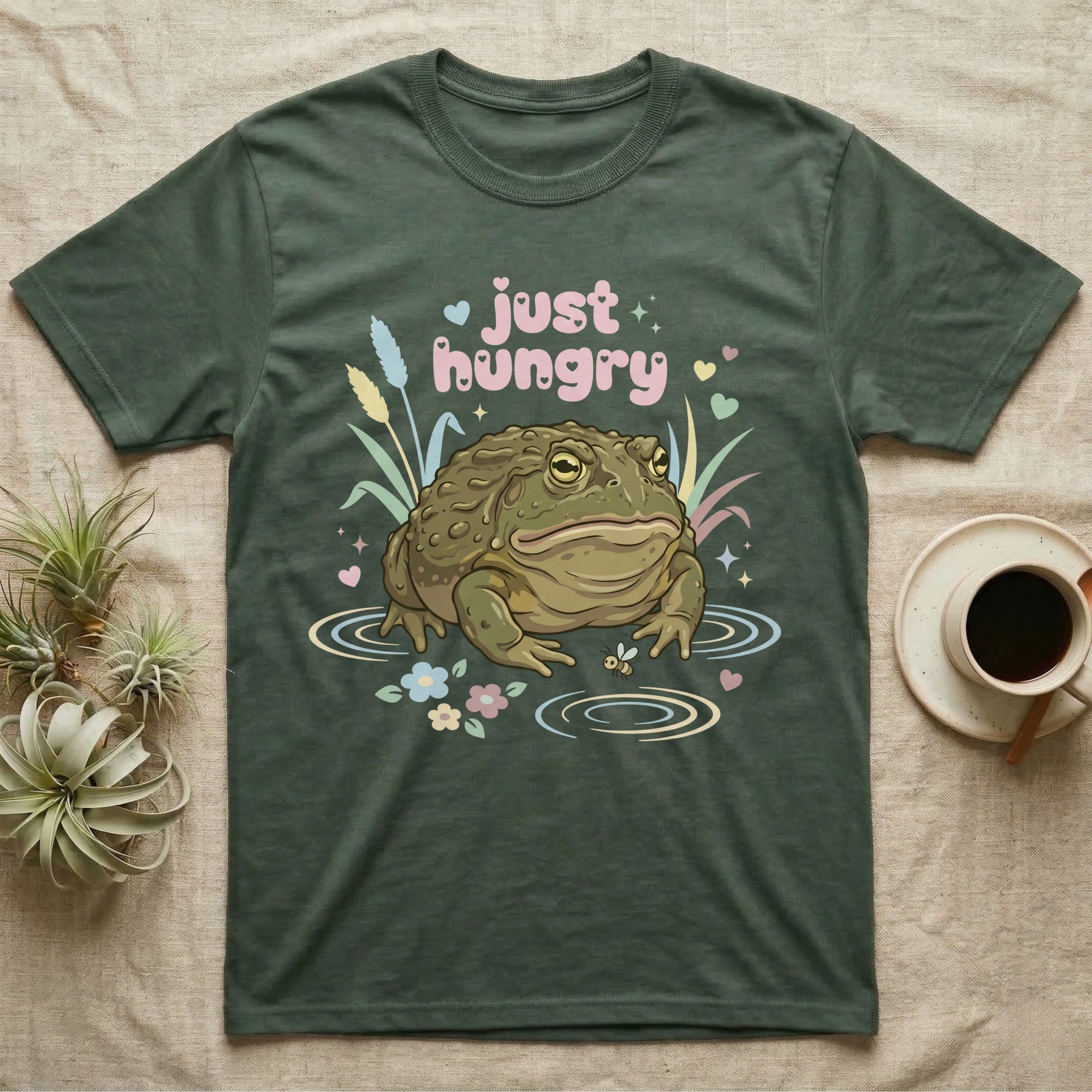 African Bullfrog Baby T-Shirt in Military Green — Pastel African bullfrog eyeing a tiny bug among reeds and flowers, puffy pink bubble wordmark "just hungry" above.