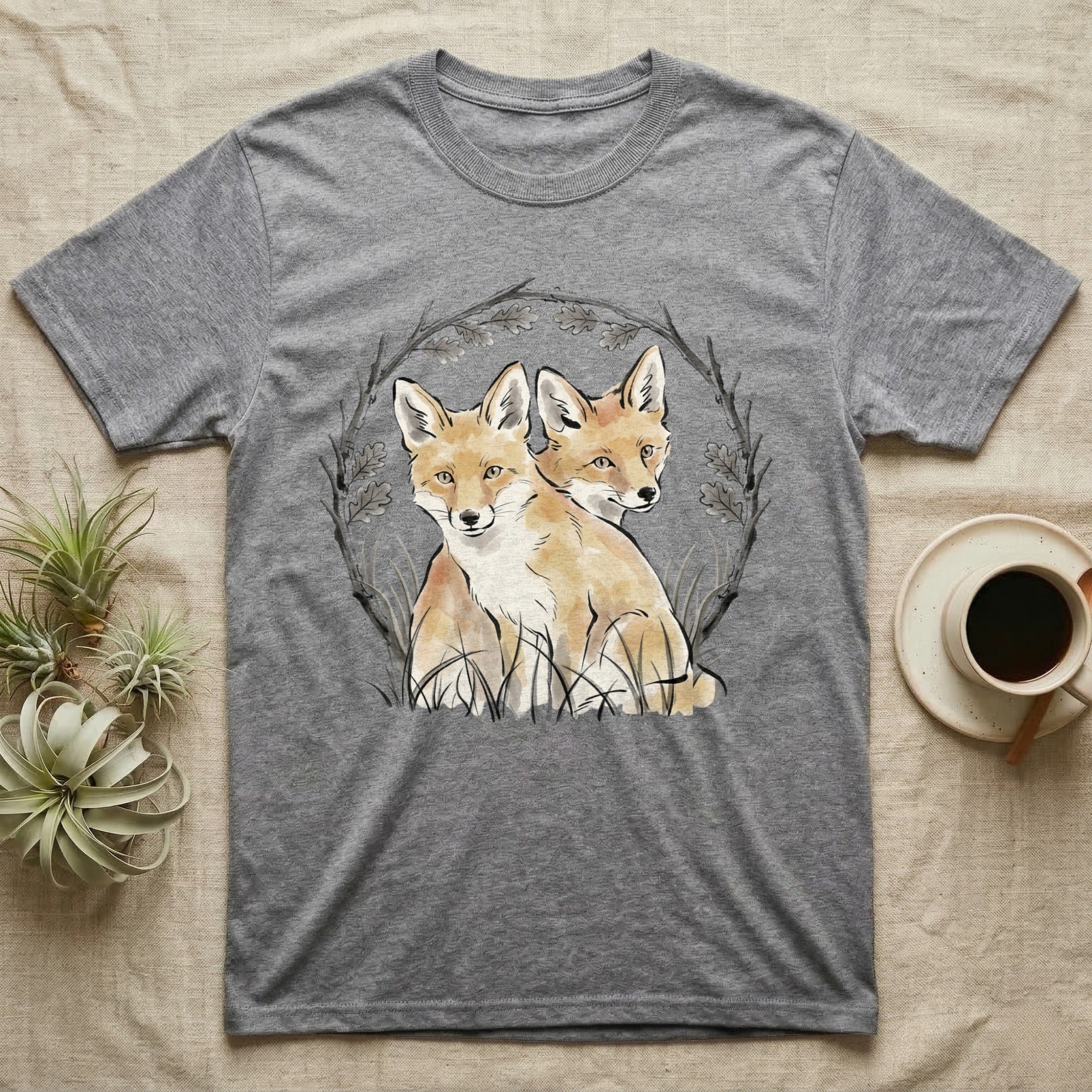 Two foxes sit in a circular arrangement of branches and leaves on a grey t-shirt.