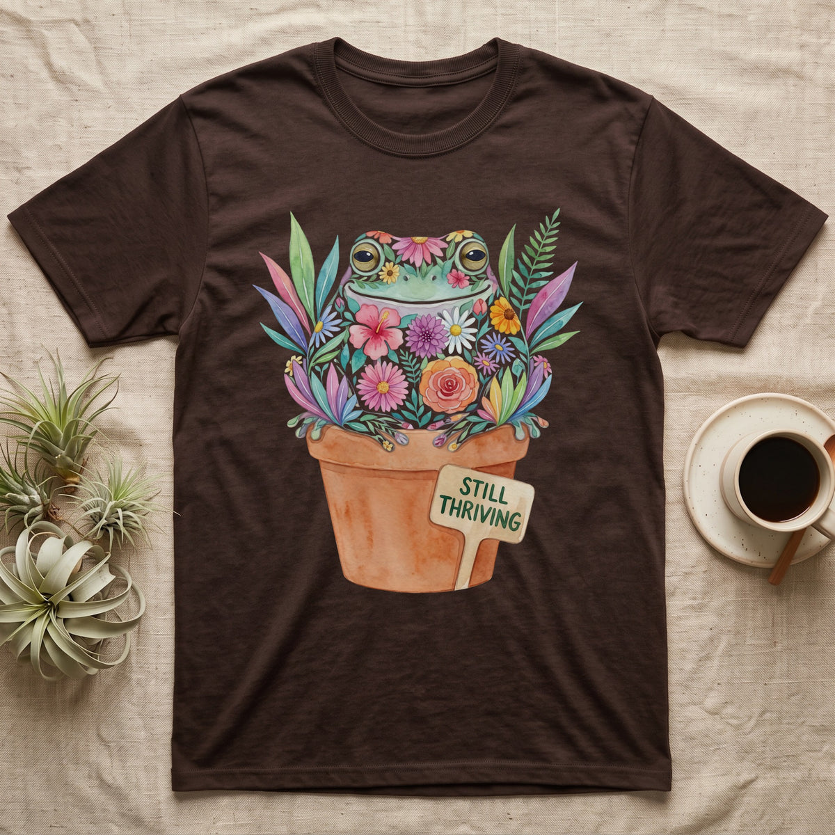 Tree Frog Thriving T-Shirt in Dark Chocolate — A White's tree frog made of watercolour flowers, sitting in a terracotta pot with a plant label reading STILL THRIVING.