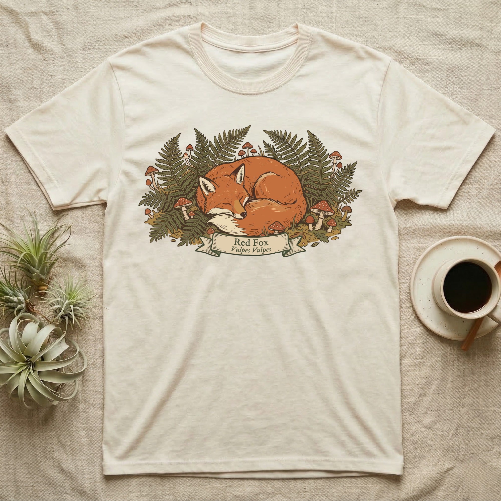 Cream t-shirt with a sleeping fox surrounded by ferns and mushrooms.