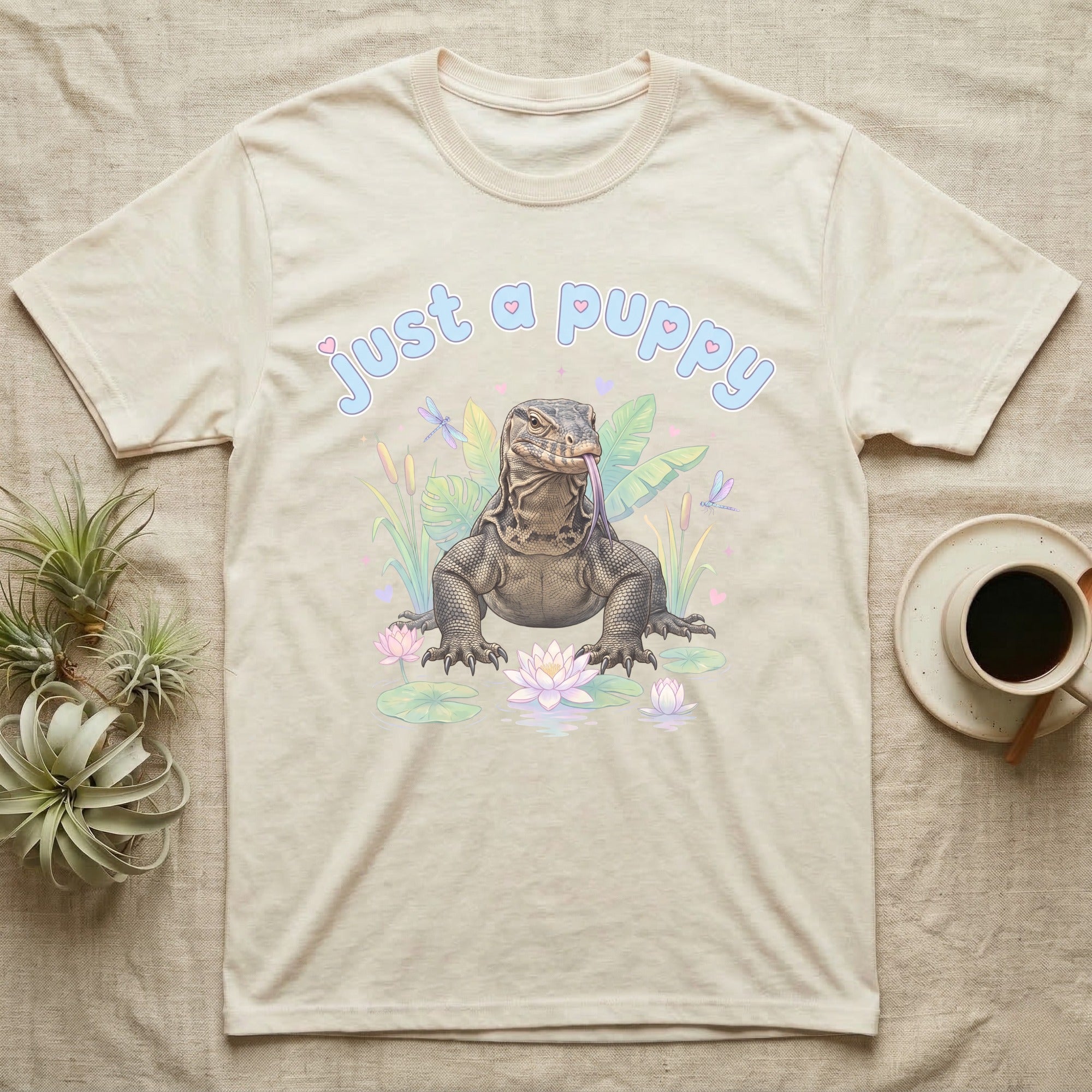 Asian Water Monitor Baby T-Shirt in Natural — Asian water monitor with tongue out among pastel reeds and lotus under puffy blue bubble text "just a puppy".