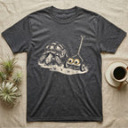 Tortoise Lawnmower Googly T-Shirt in Dark Heather — Linocut illustration of a sulcata tortoise grazing beside a push mower wearing oversized googly eyes.