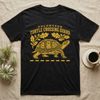Box Turtle Crossing T-Shirt in Black — Yellow folk-print eastern box turtle walking across a dashed road line with oak leaves, text Volunteer Turtle Crossing Guard