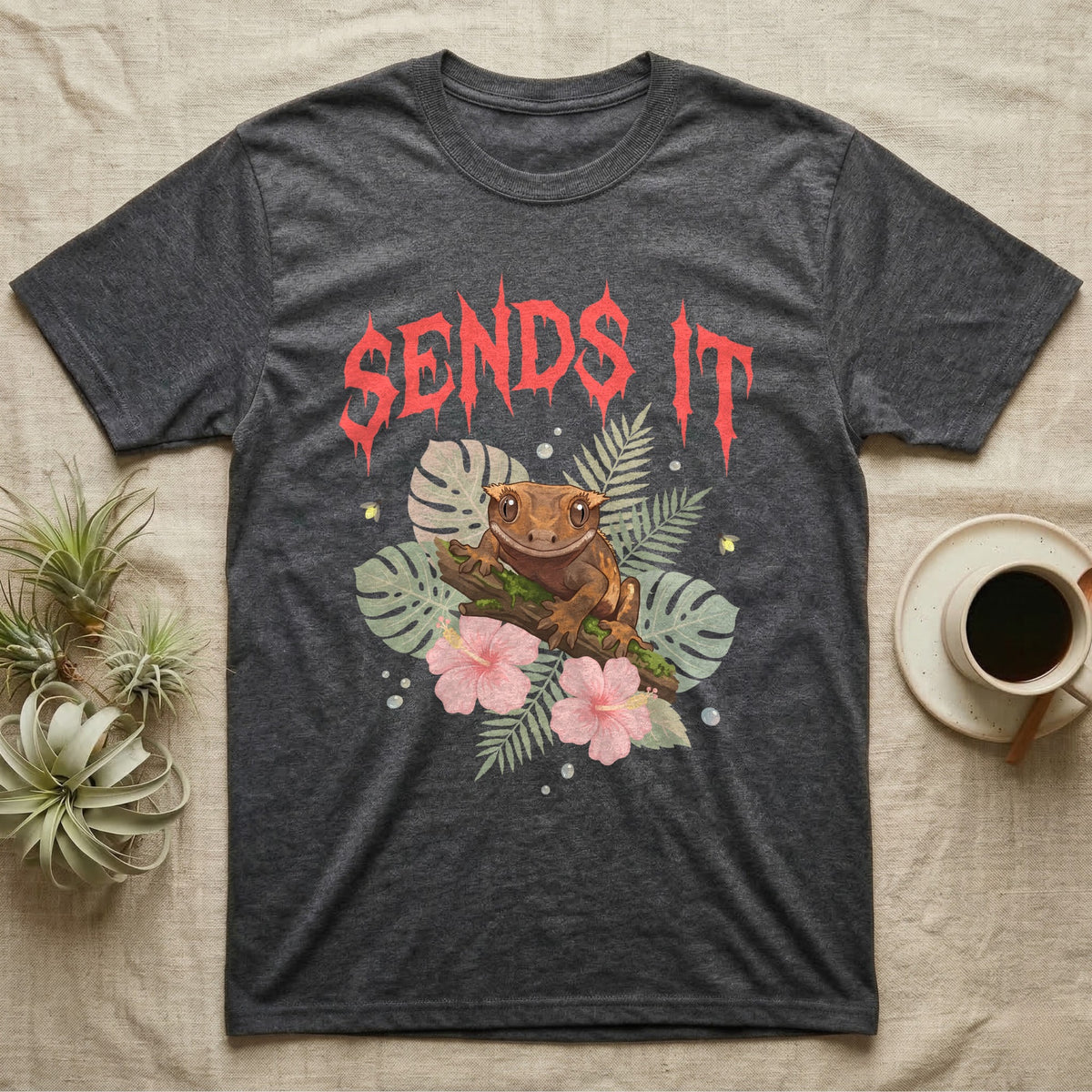 Crested Gecko Feral T-Shirt in Dark Heather — Crested gecko on a mossy branch in a pastel jungle scene under a spiky coral metal wordmark reading SENDS IT.