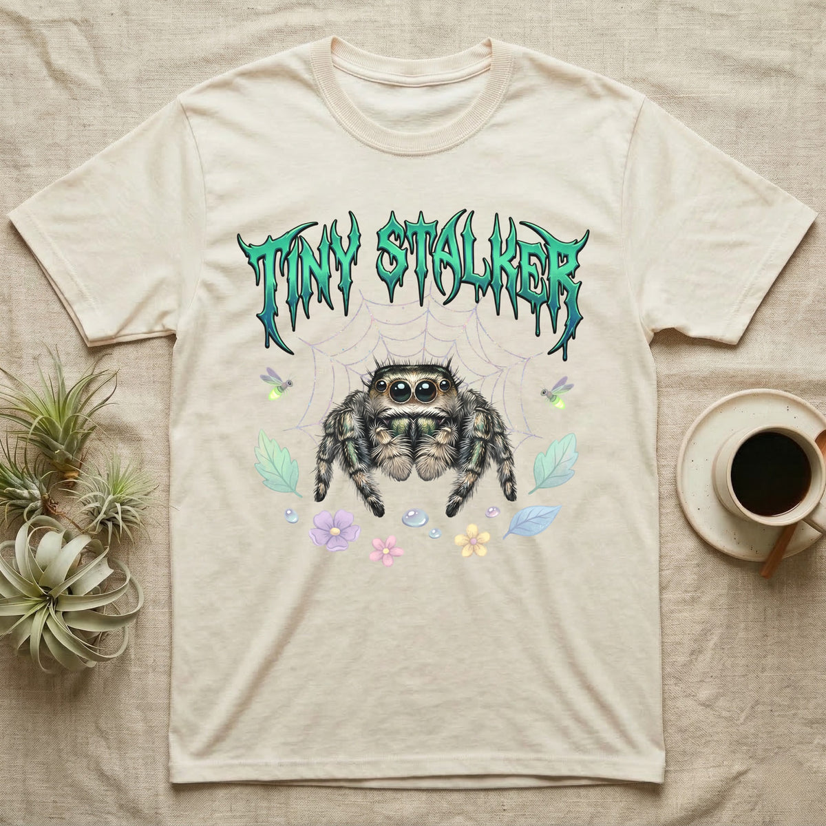 Regal Jumping Spider Feral T-Shirt in Natural — Fuzzy jumping spider with huge eyes in a pastel garden under a spiky iridescent-teal metal wordmark reading TINY STALKER.