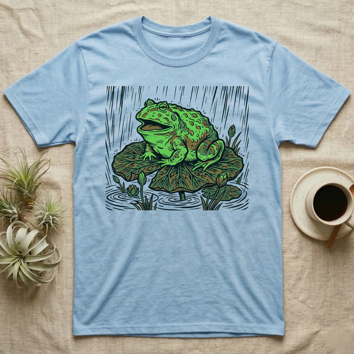 Light blue t-shirt with graphic of a frog on a lily pad in the rain.