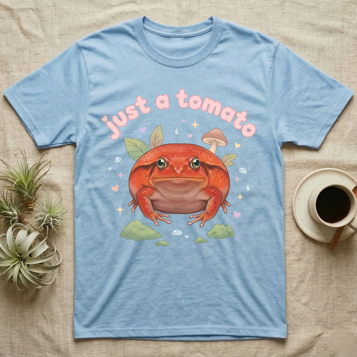 Tomato Frog Softie T-Shirt in Light Blue — Round red-orange tomato frog facing forward under a puffy pastel "just a tomato" bubble wordmark with tiny hearts.