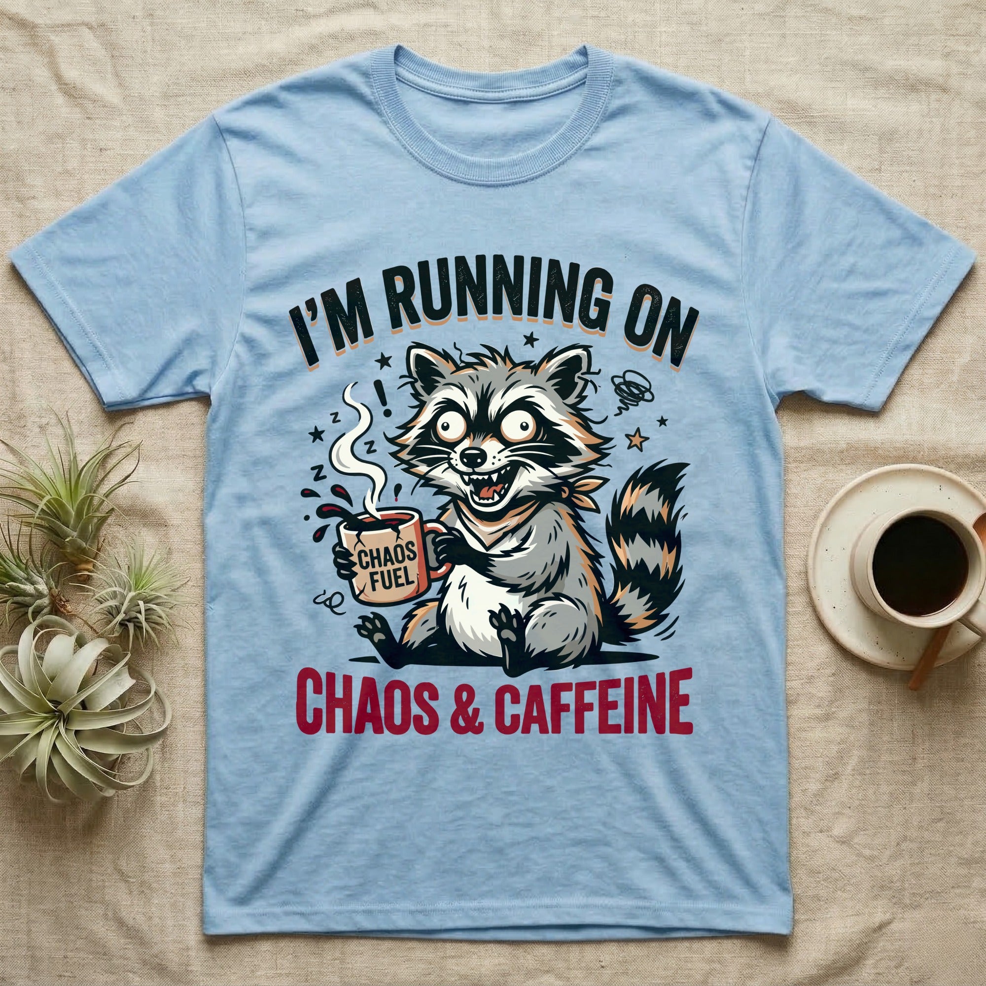 T-shirt graphic: Raccoon holding a coffee cup that says "Chaos Fuel," text reads "I'm Running On Chaos & Caffeine."