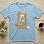 Light blue t-shirt with a framed illustration of a chameleon in a terrarium.