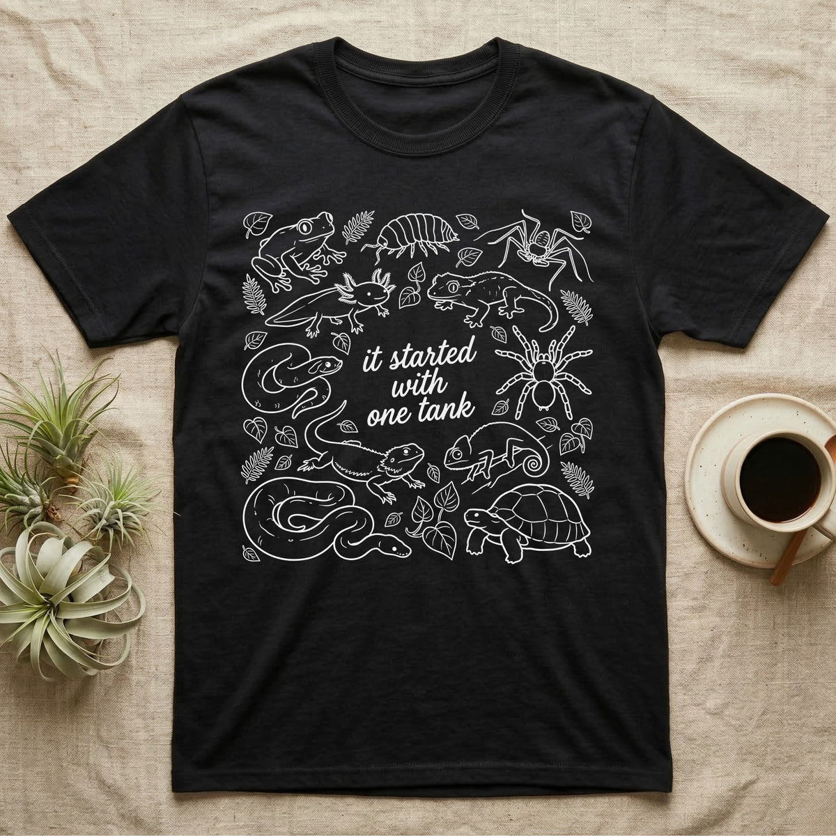 Terrarium Keeper One Tank T-Shirt in Black — White outline illustration of eleven terrarium species — bearded dragon, crested gecko, ball python, axolotl, tortoise, tarantula, isopod, whip spider, chameleon, tree frog and hognose — with pothos and fern leaves, ringed around the script line it started with one tank.