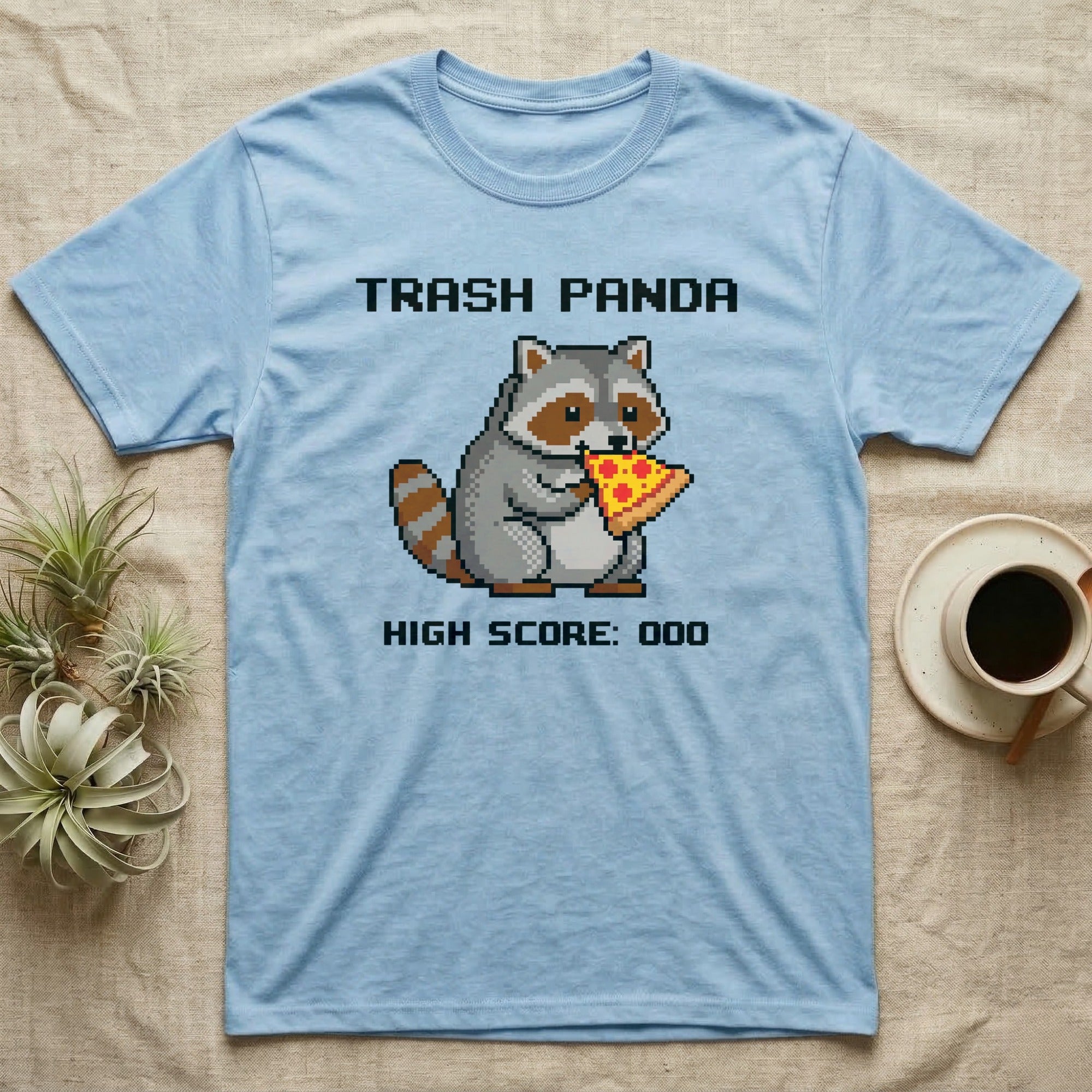 Light blue t-shirt with pixelated raccoon eating pizza and text "Trash Panda High Score: 000".