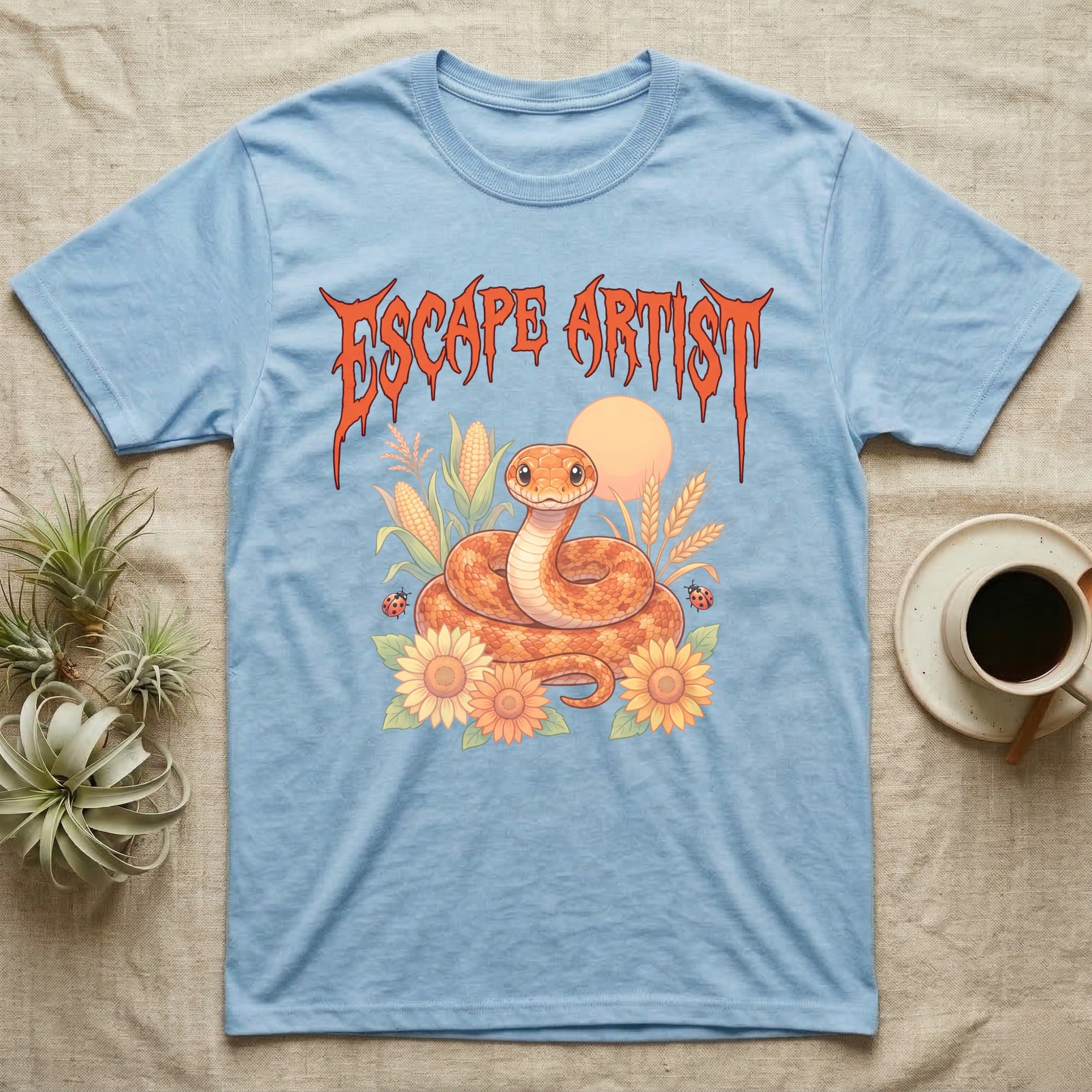 Corn Snake Feral T-Shirt in Light Blue — Orange corn snake coiled in a pastel cornfield scene under a spiky blood-orange metal wordmark reading ESCAPE ARTIST.