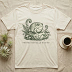 Cream t-shirt with a toad illustration on a rock surrounded by plants and mushrooms.