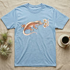 Crested Gecko Blossom T-Shirt in Light Blue — Watercolor illustration of a rust-brown crested gecko holding a frangipani blossom sprig, on a dark grey t-shirt.