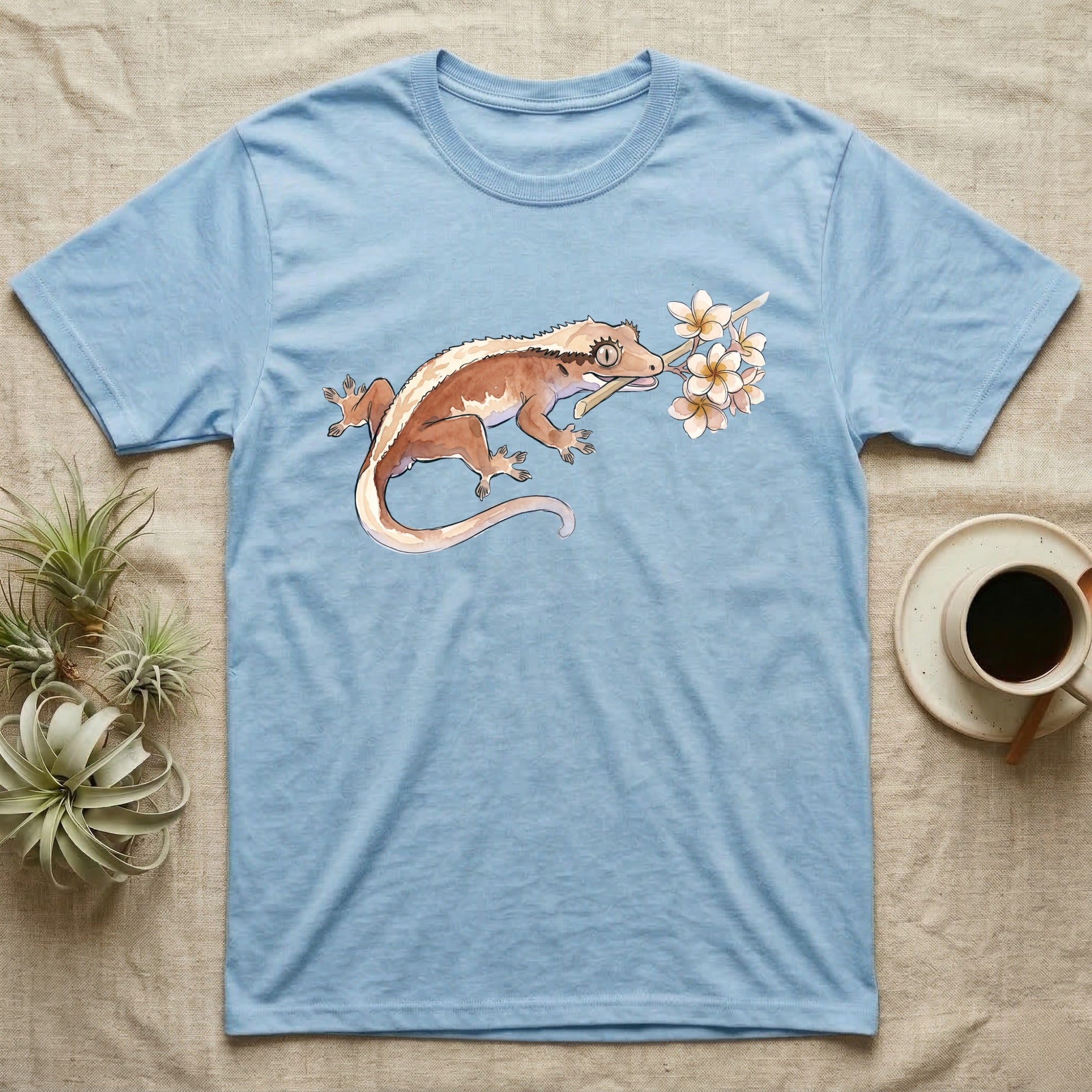 Crested Gecko Blossom T-Shirt in Light Blue — Watercolor illustration of a rust-brown crested gecko holding a frangipani blossom sprig, on a dark grey t-shirt.