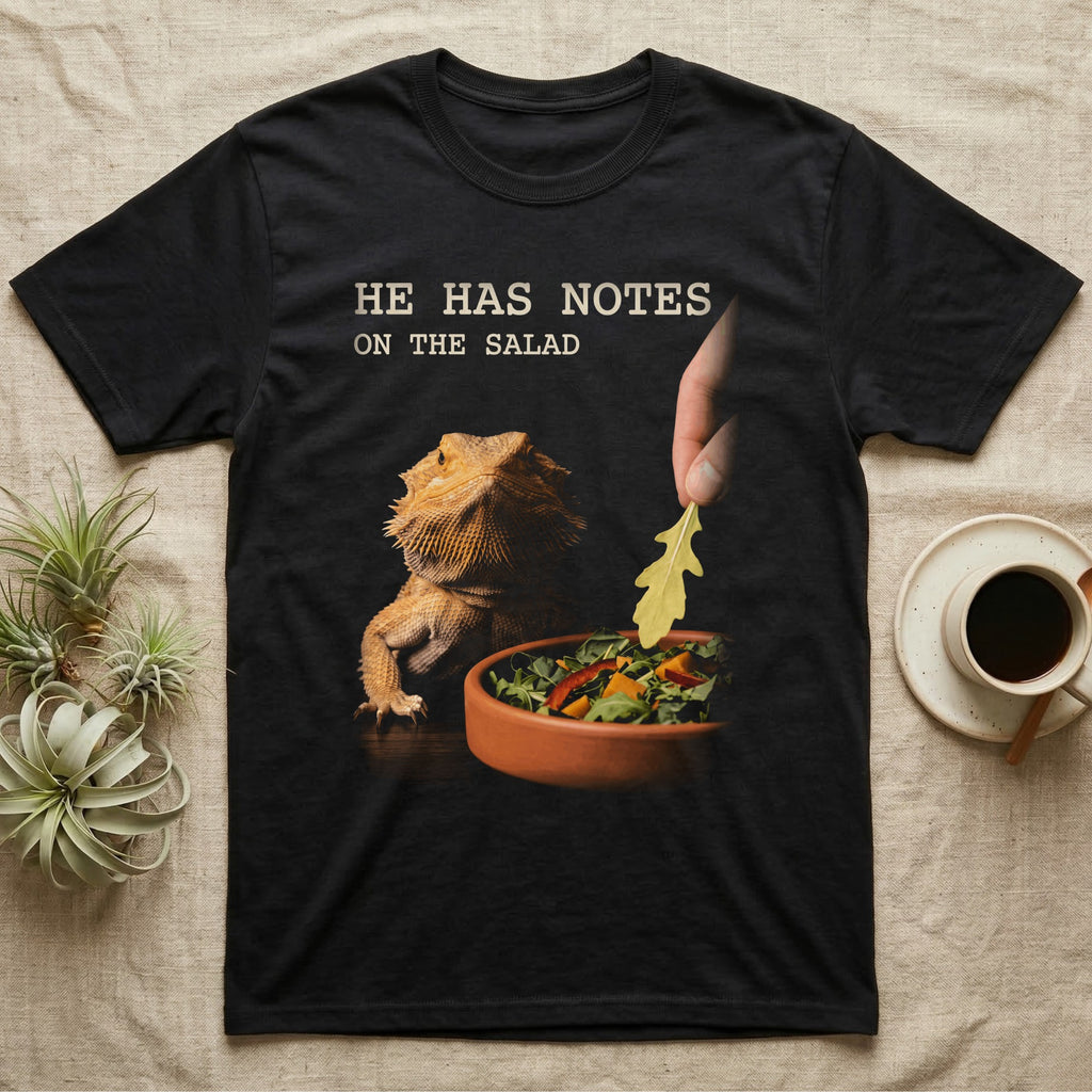 Bearded Dragon Salad Notes T-Shirt in Black — Photorealistic bearded dragon staring flatly as a hand lowers an arugula leaf into a foreground salad dish.