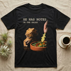 Bearded Dragon Salad Notes T-Shirt in Black — Photorealistic bearded dragon staring flatly as a hand lowers an arugula leaf into a foreground salad dish.