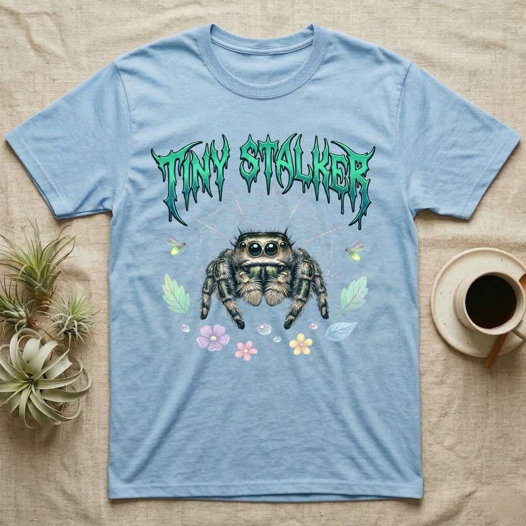 Regal Jumping Spider Feral T-Shirt in Light Blue — Fuzzy jumping spider with huge eyes in a pastel garden under a spiky iridescent-teal metal wordmark reading TINY STALKER.