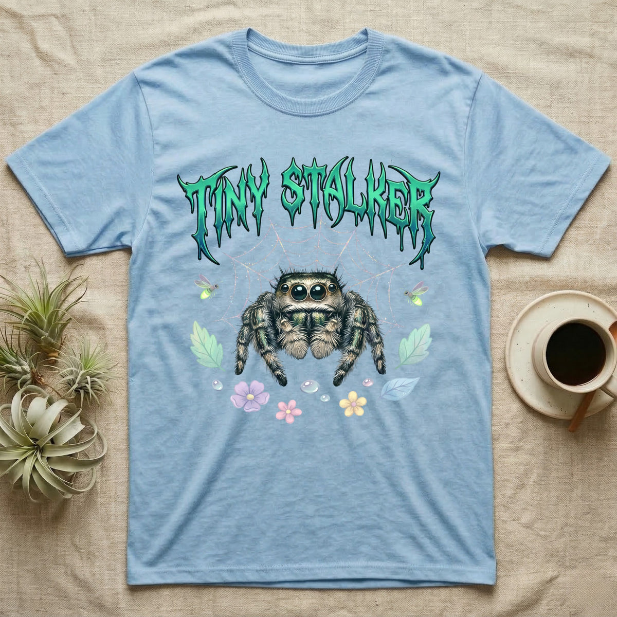 Regal Jumping Spider Feral T-Shirt in Light Blue — Fuzzy jumping spider with huge eyes in a pastel garden under a spiky iridescent-teal metal wordmark reading TINY STALKER.