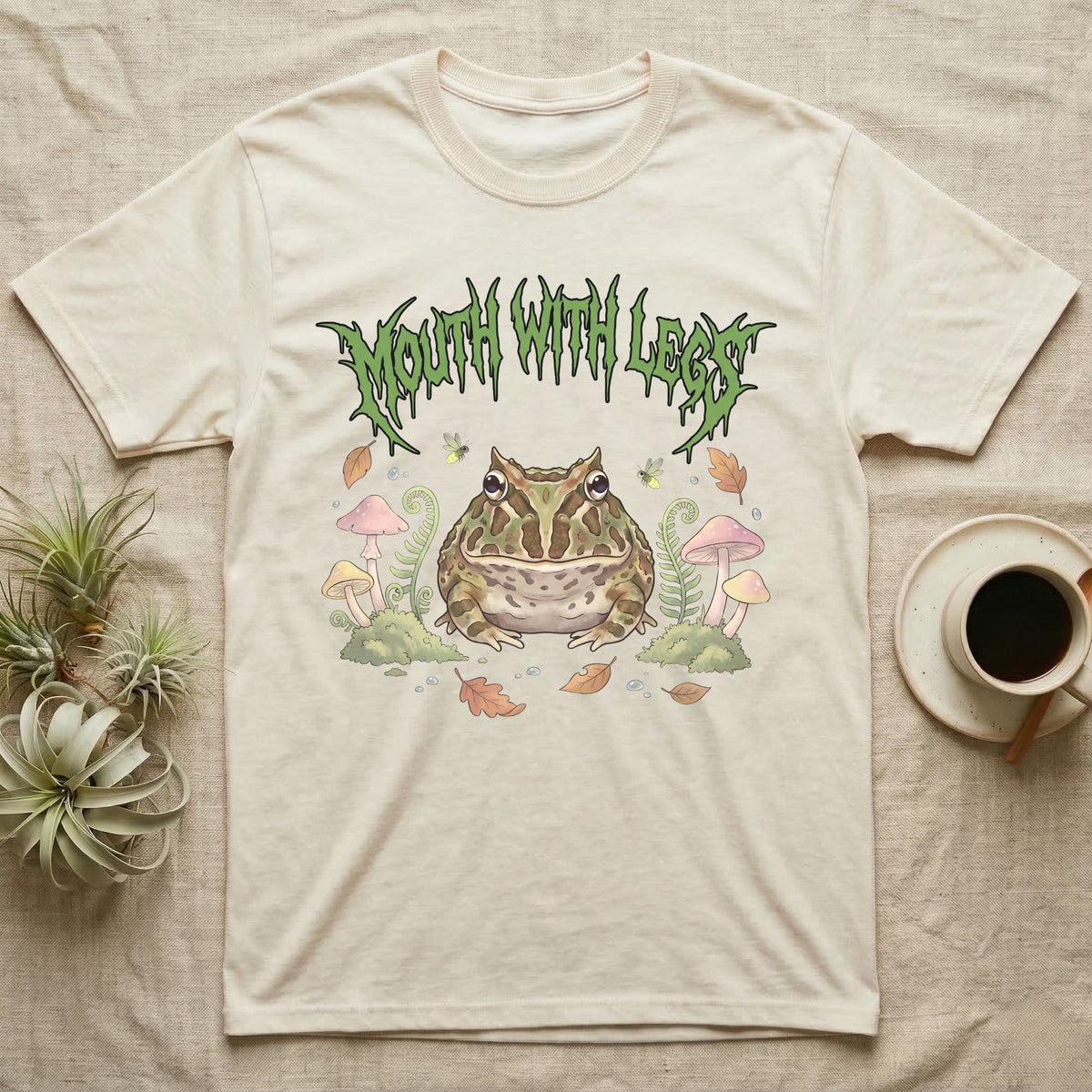 Pacman Frog Feral T-Shirt in Natural — Round Pacman frog in a pastel forest-floor scene under a spiky leaf-green metal wordmark reading MOUTH WITH LEGS.