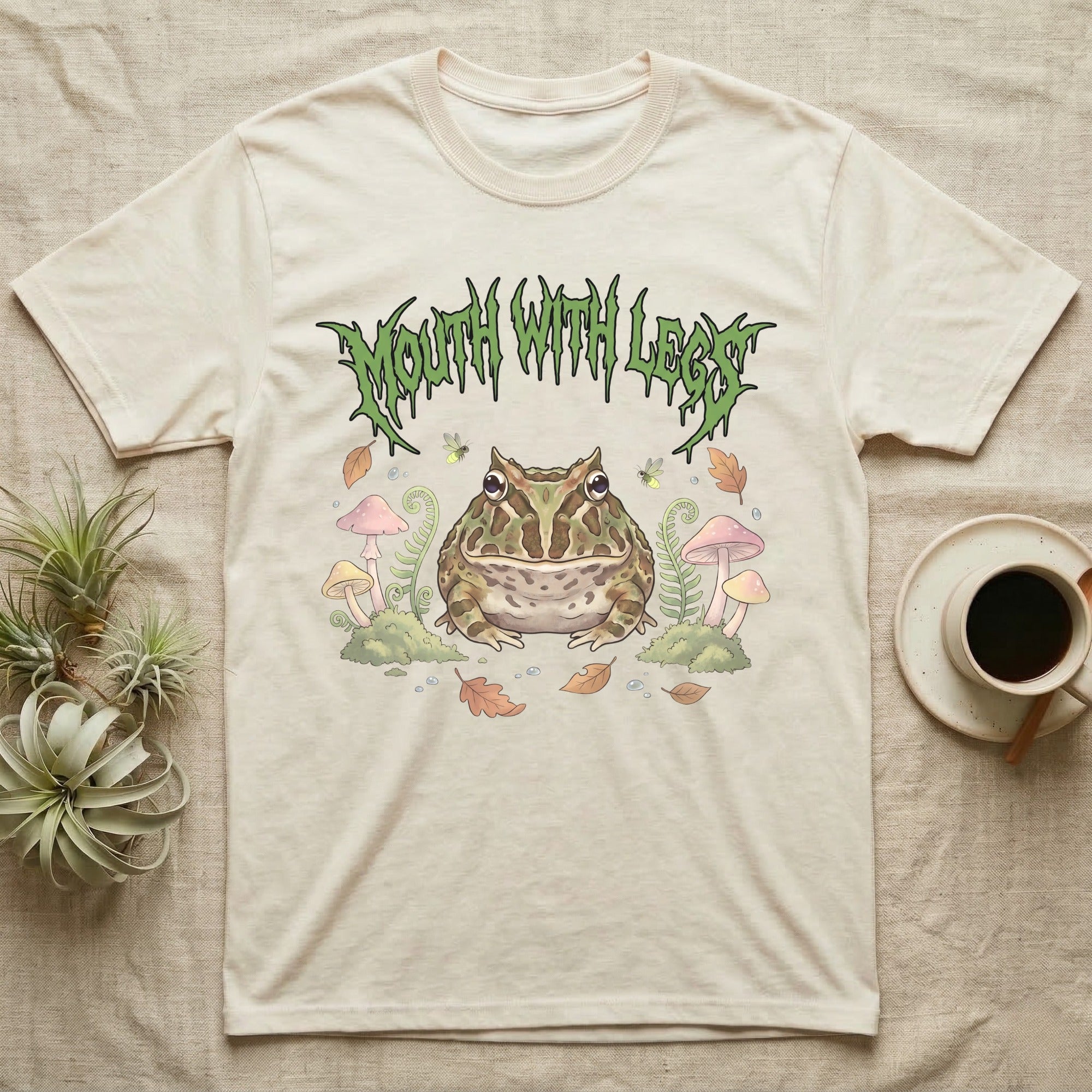 Pacman Frog Feral T-Shirt in Natural — Round Pacman frog in a pastel forest-floor scene under a spiky leaf-green metal wordmark reading MOUTH WITH LEGS.