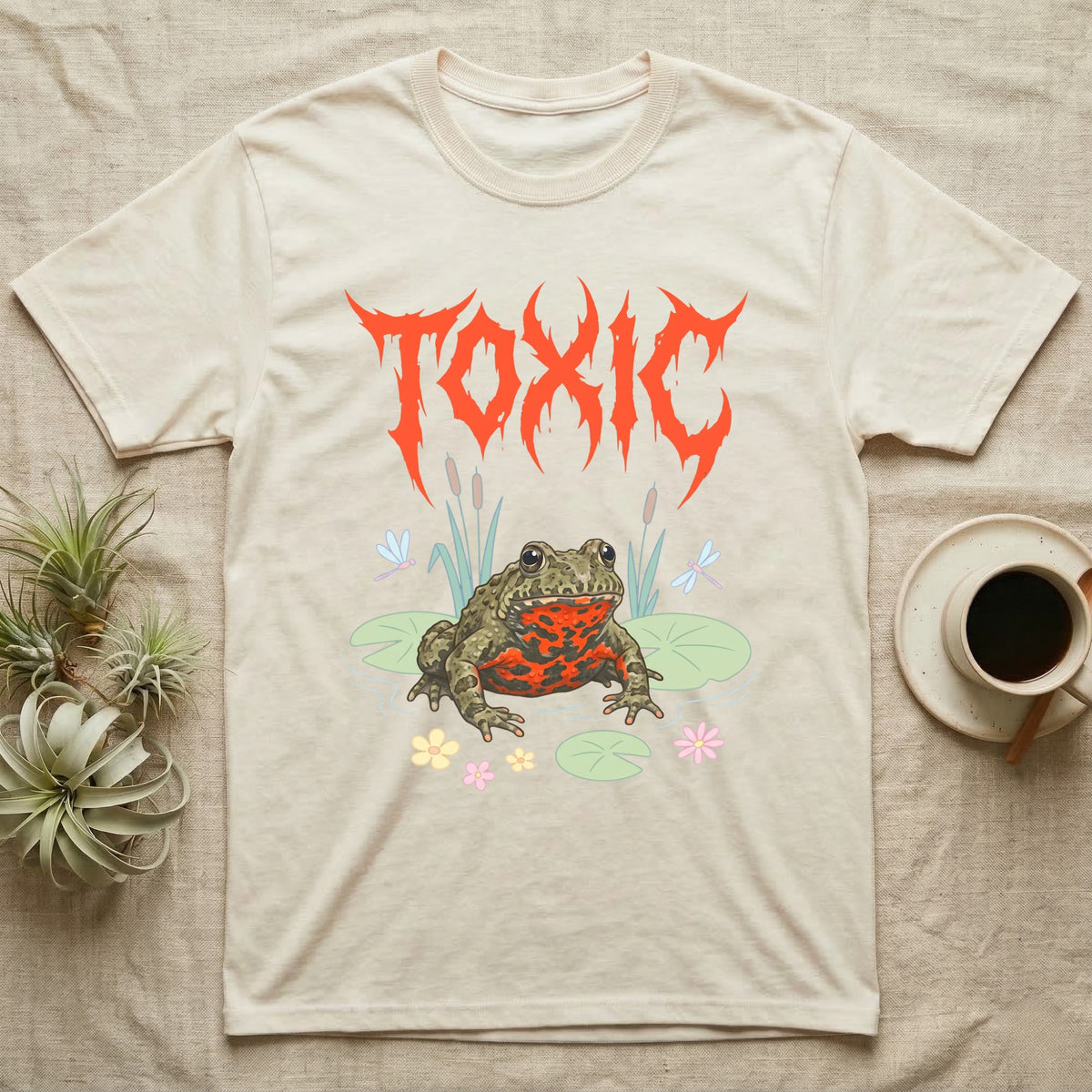 Oriental Fire-Bellied Toad Feral T-Shirt in Natural — Pastel fire-bellied toad with red-orange belly among lily pads and dragonflies, jagged metal wordmark "TOXIC" above.