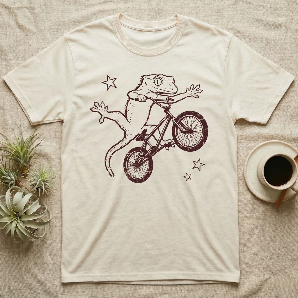 Crested Gecko BMX T-Shirt in Natural — Distressed one-ink oxblood screenprint of a crested gecko airborne on a BMX bike mid-barspin, front hands thrown wide, back feet gripping the pedals.