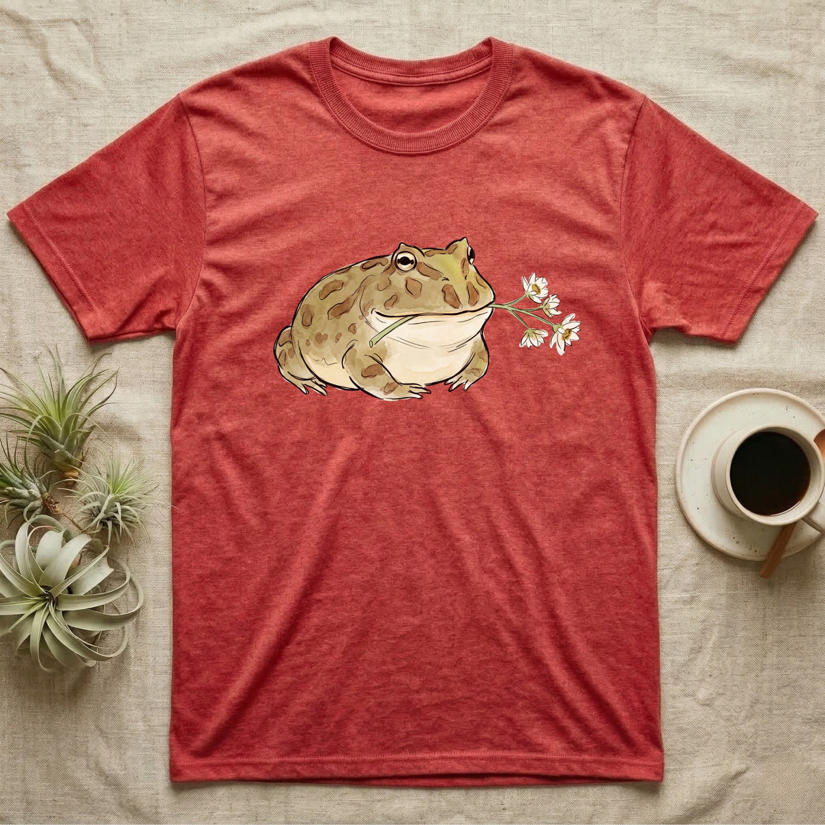 Pacman Frog Blossom T-Shirt in Heather Red — Watercolor illustration of an olive-tan Pacman frog holding a water-lily blossom sprig, on a dark grey t-shirt.