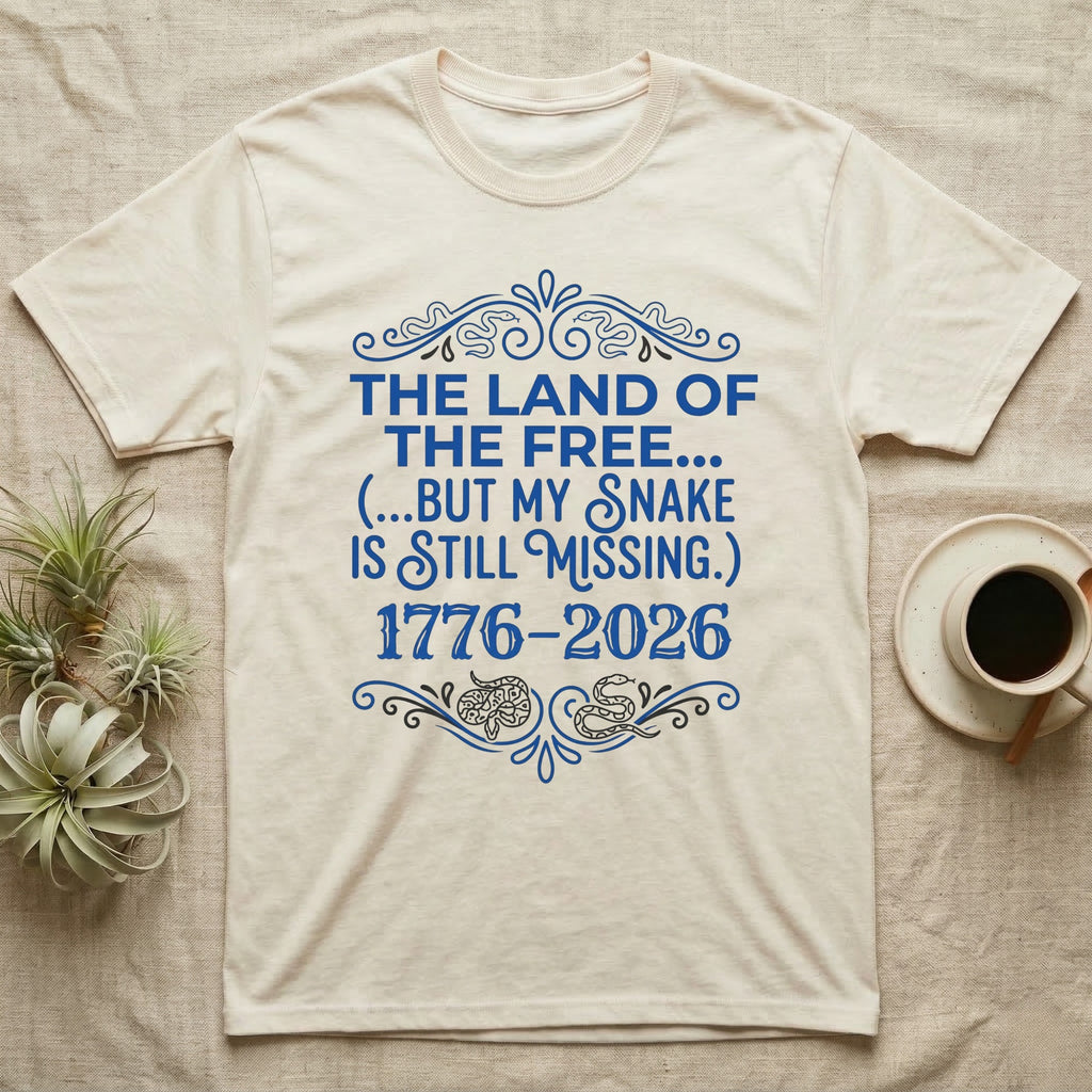Corn Snake Still Missing Tee in Natural — Royal blue typographic tee reading The land of the free but my snake is still missing, with Victorian filigree and small coiled corn snakes.