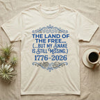 Corn Snake Still Missing Tee in Natural — Royal blue typographic tee reading The land of the free but my snake is still missing, with Victorian filigree and small coiled corn snakes.
