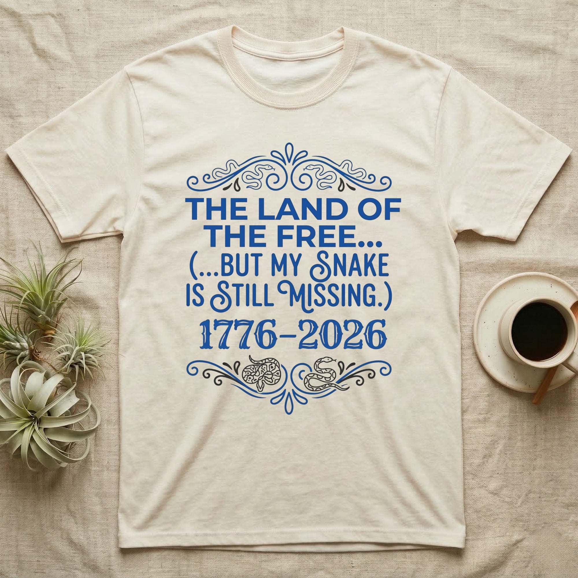 Corn Snake Still Missing Tee in Natural — Royal blue typographic tee reading The land of the free but my snake is still missing, with Victorian filigree and small coiled corn snakes.