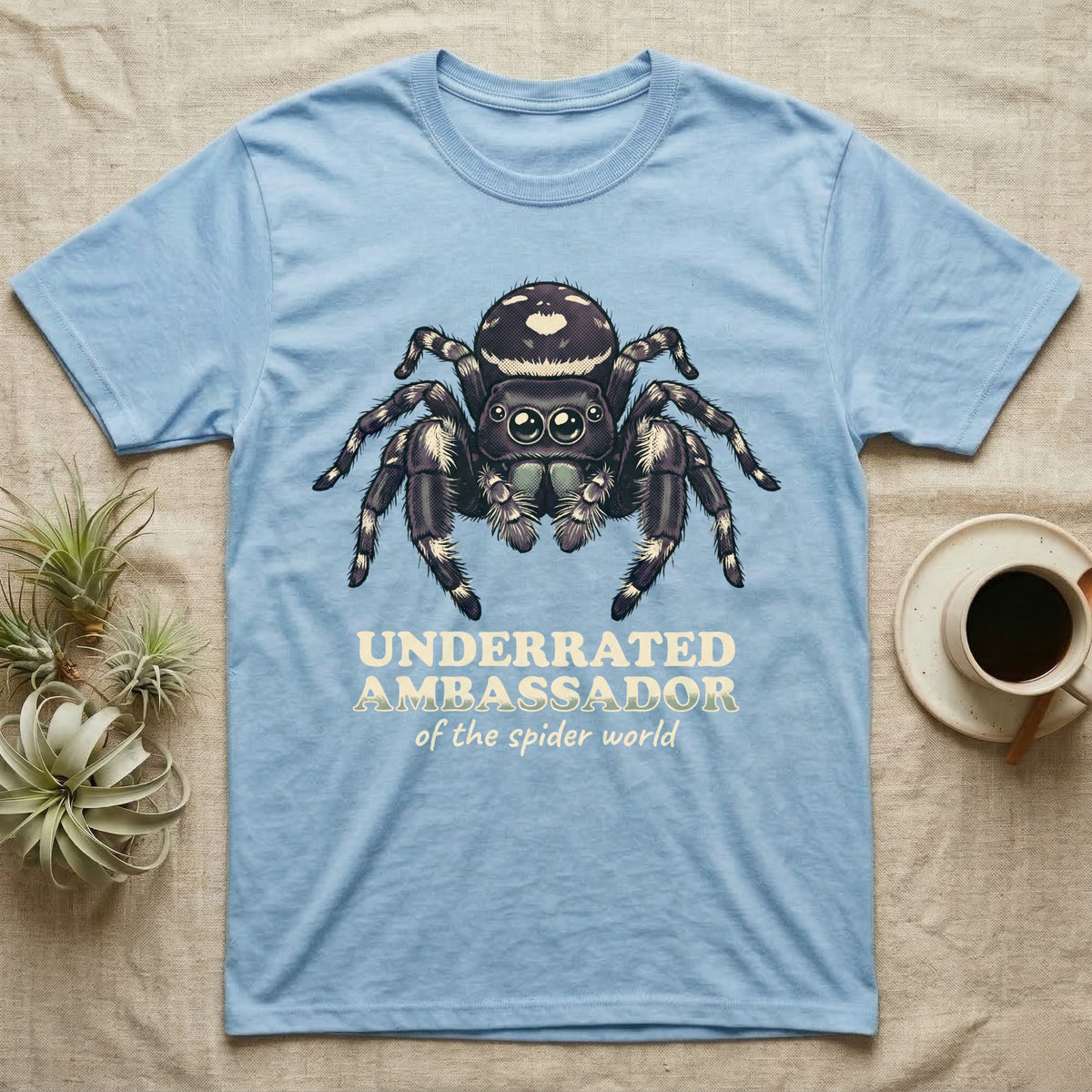 Underrated Ambassador Spider T-Shirt in Light Blue — Soft creepy-cute portrait of a big-eyed jumping spider with hand-lettered text reading Underrated Ambassador of the Spider World.