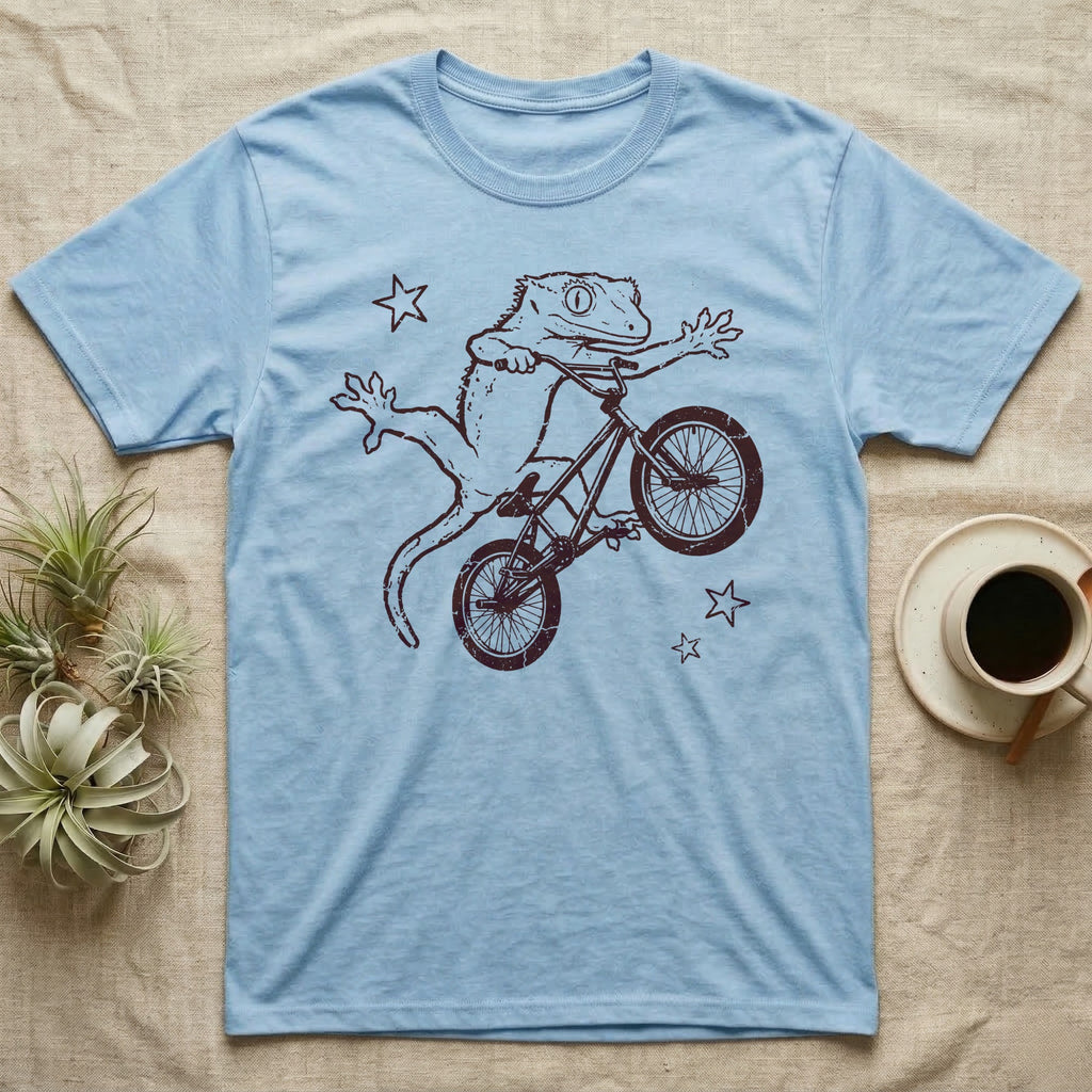 Crested Gecko BMX T-Shirt in Light Blue — Distressed one-ink oxblood screenprint of a crested gecko airborne on a BMX bike mid-barspin, front hands thrown wide, back feet gripping the pedals.