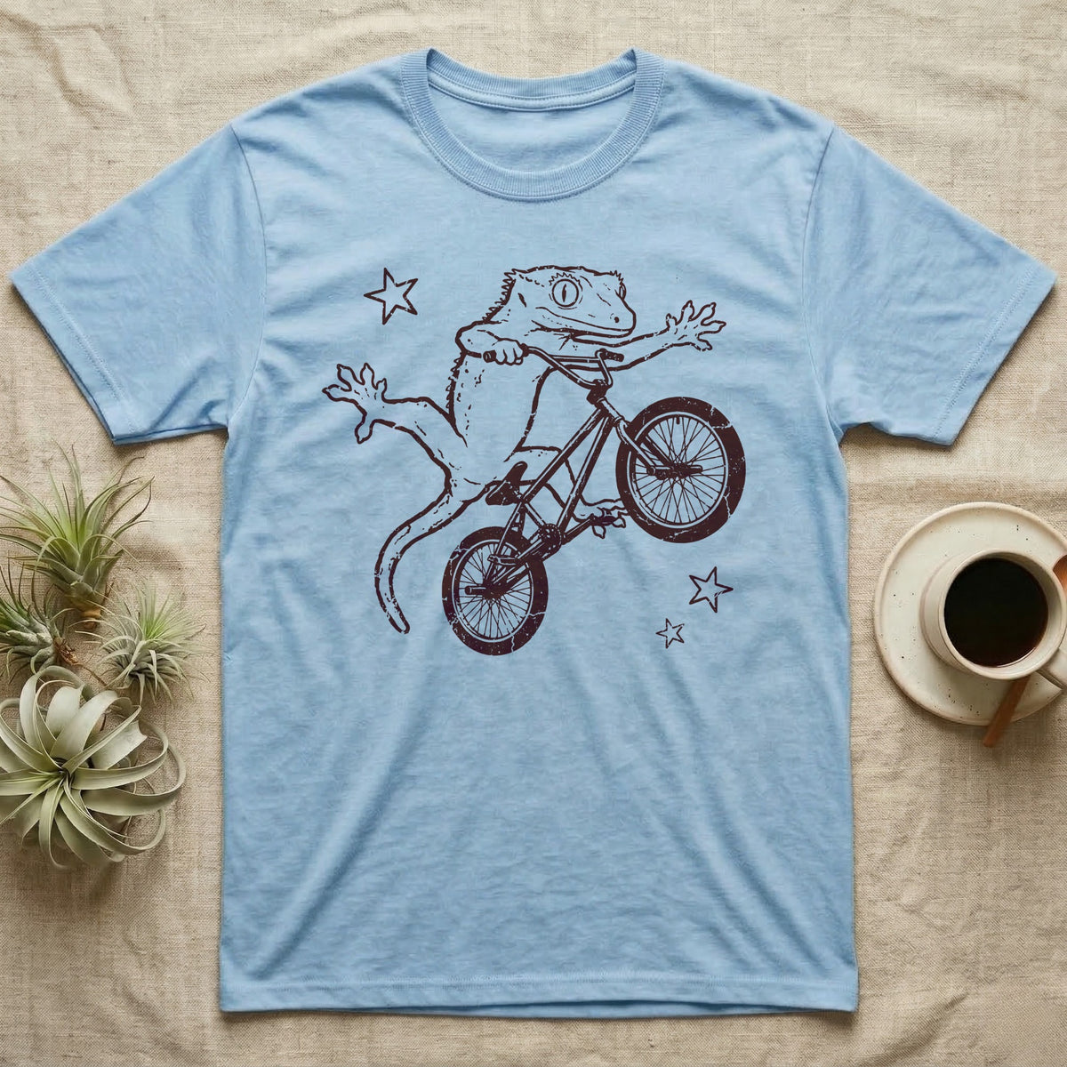 Crested Gecko BMX T-Shirt in Light Blue — Distressed one-ink oxblood screenprint of a crested gecko airborne on a BMX bike mid-barspin, front hands thrown wide, back feet gripping the pedals.