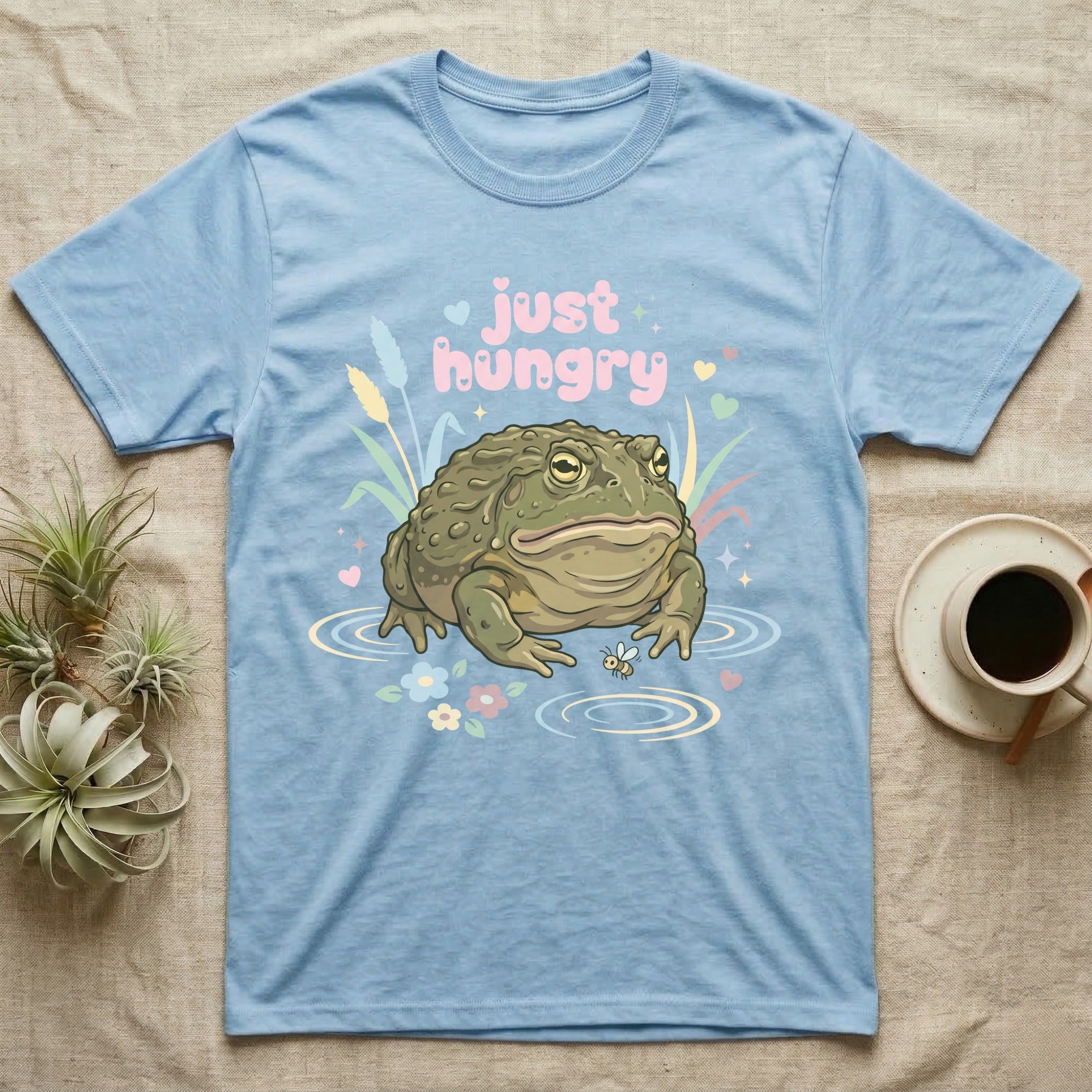 African Bullfrog Baby T-Shirt in Light Blue — Pastel African bullfrog eyeing a tiny bug among reeds and flowers, puffy pink bubble wordmark "just hungry" above.