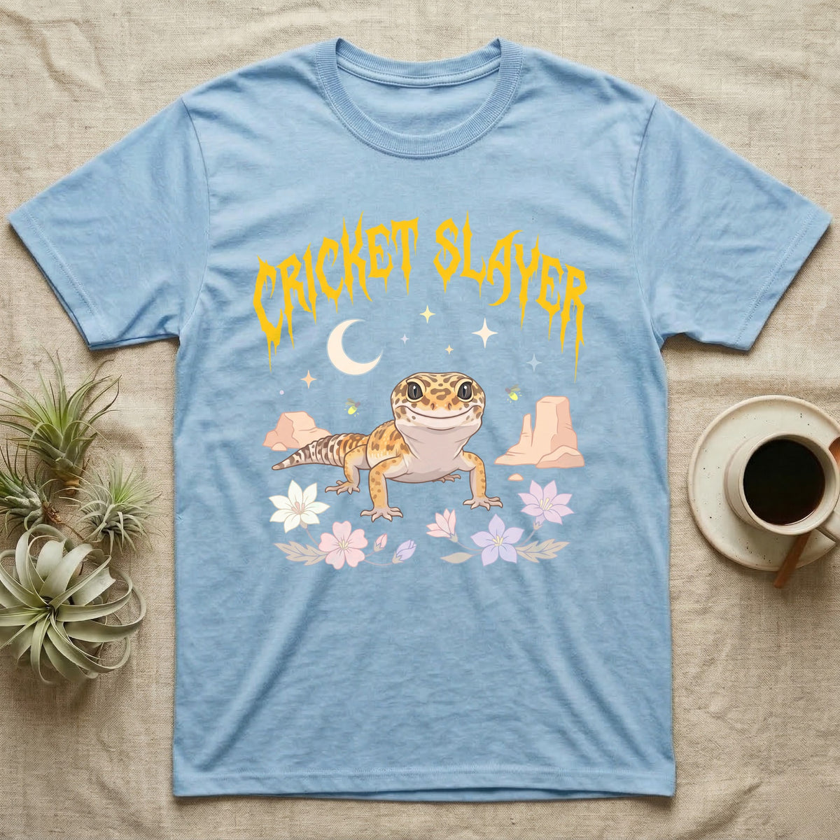 Leopard Gecko Feral T-Shirt in Light Blue — Smiling leopard gecko in a pastel desert-night scene under a spiky golden metal wordmark reading CRICKET SLAYER.