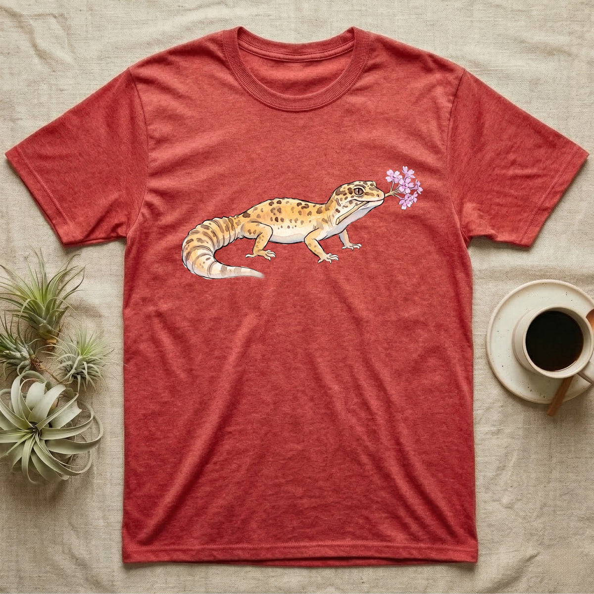 Leopard Gecko Blossom T-Shirt in Heather Red — Watercolor illustration of a sandy-gold spotted leopard gecko holding a sand verbena blossom sprig, on a dark grey t-shirt.