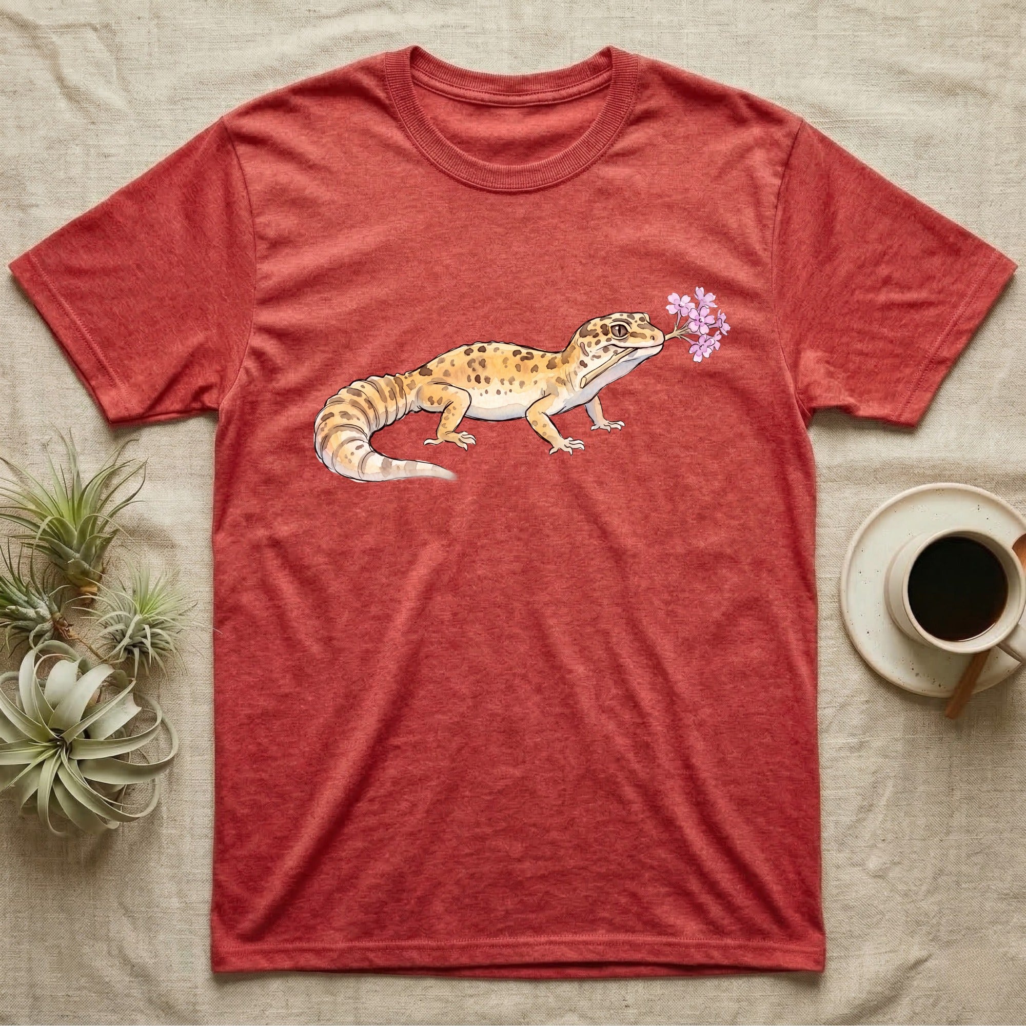 Leopard Gecko Blossom T-Shirt in Heather Red — Watercolor illustration of a sandy-gold spotted leopard gecko holding a sand verbena blossom sprig, on a dark grey t-shirt.