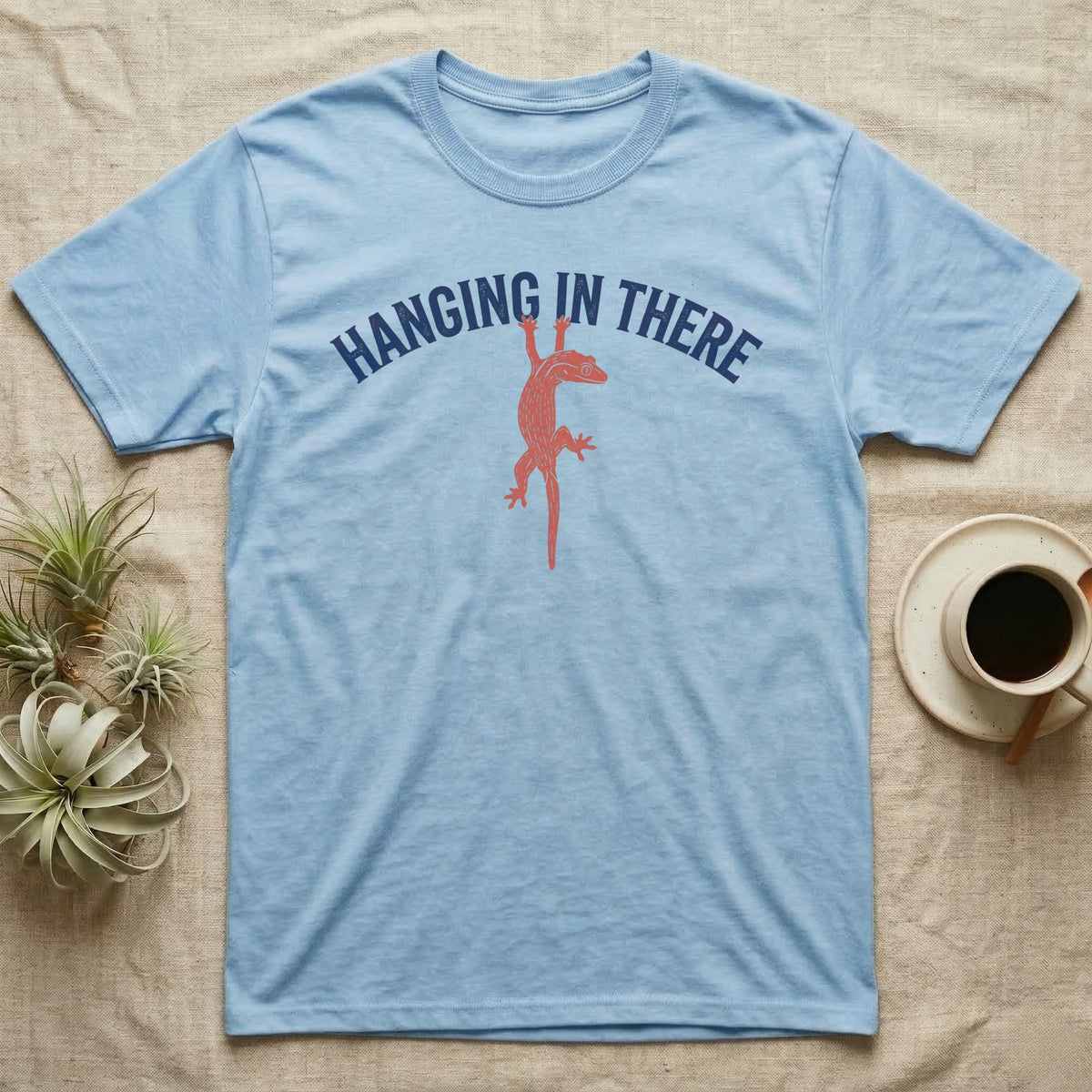 Crested Gecko Hanging T-Shirt in Light Blue — Red crested gecko hanging by its front feet from the navy arched words HANGING IN THERE