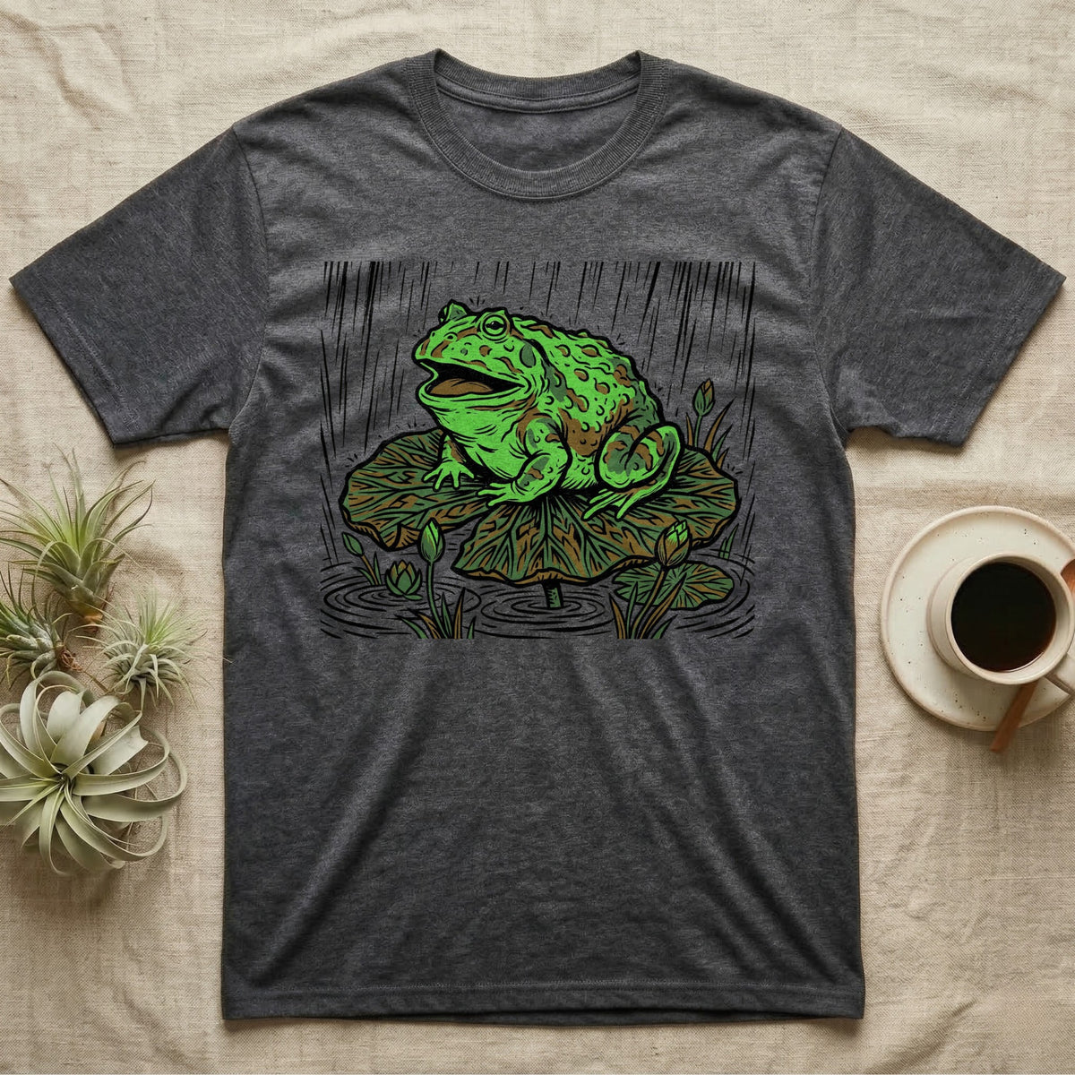 Dark grey t-shirt featuring a green frog sitting on a lily pad in the rain.