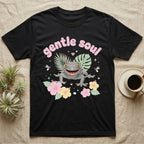 Tokay Gecko Baby T-Shirt in Black — Blue-and-orange spotted tokay gecko among pastel leaves and hibiscus under puffy pink bubble text "gentle soul".