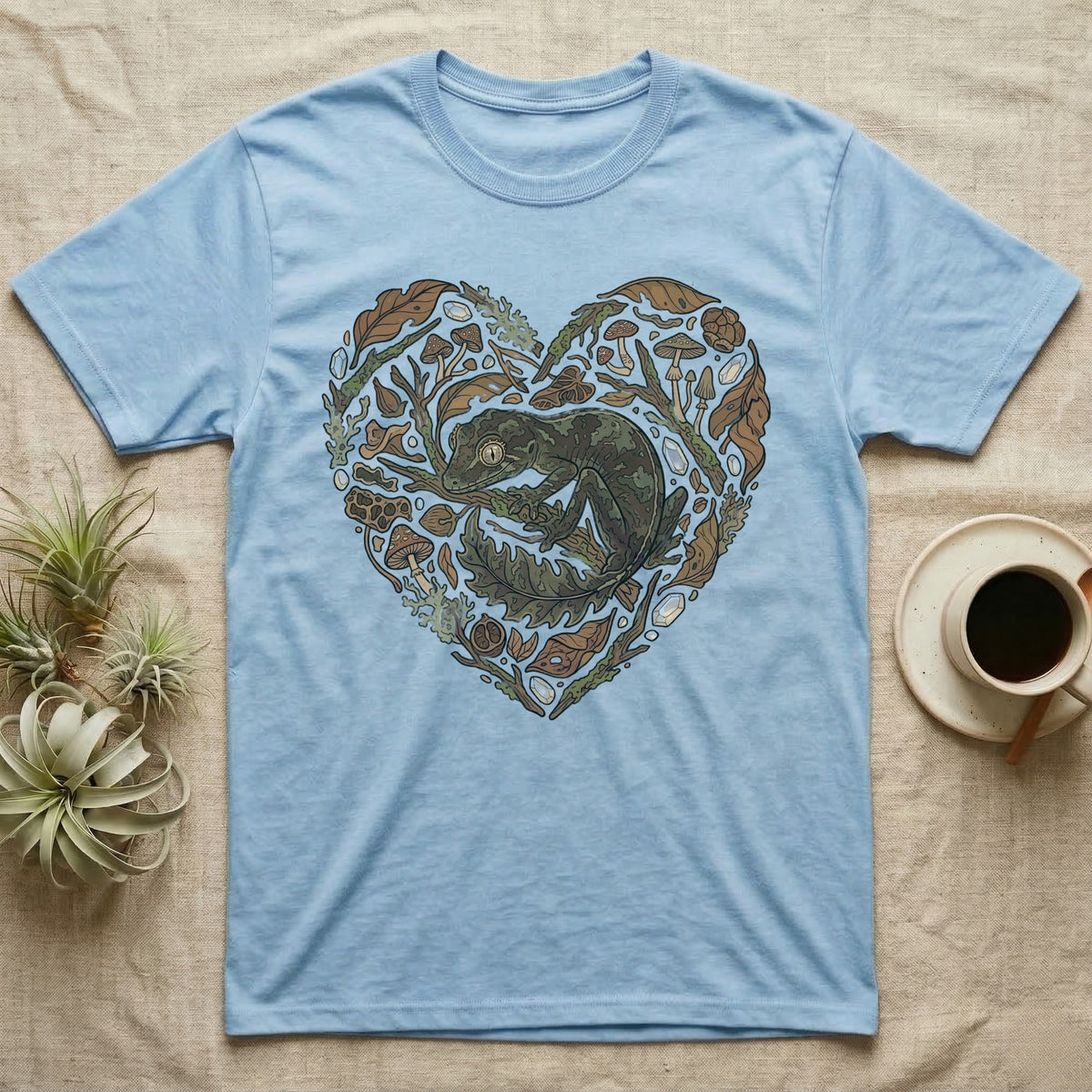 Light blue t-shirt with a green gecko in a heart shape made of leaves and mushrooms.