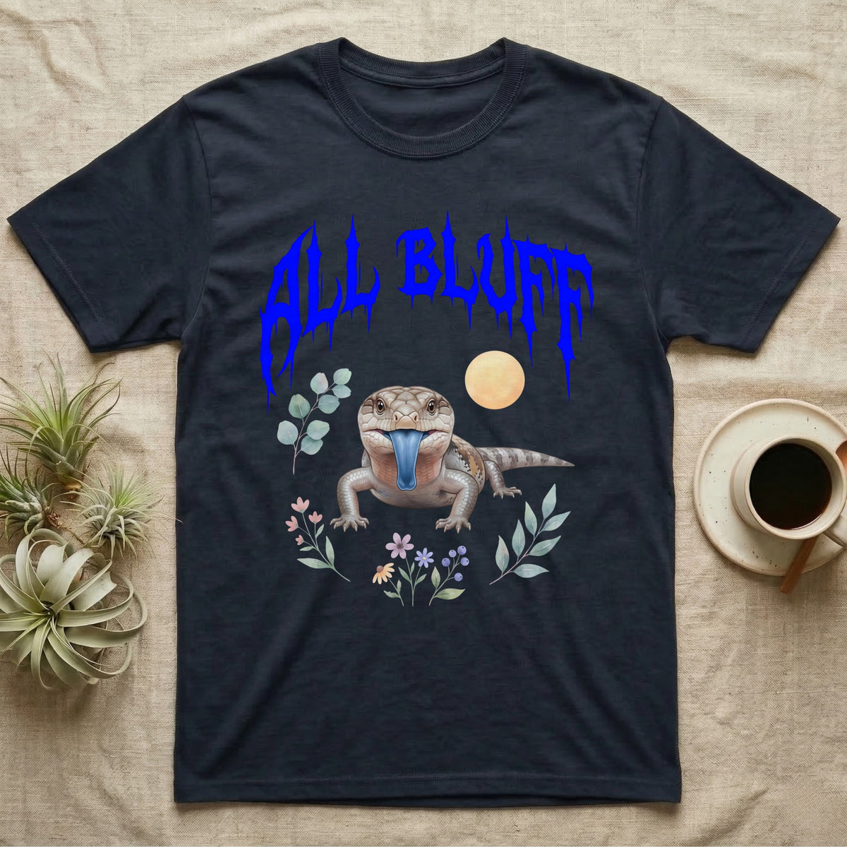 Northern Blue-Tongue Skink Feral T-Shirt in Navy — Blue-tongue skink with its blue tongue out under a jagged cobalt-blue metal "ALL BLUFF" wordmark and pastel leaves.