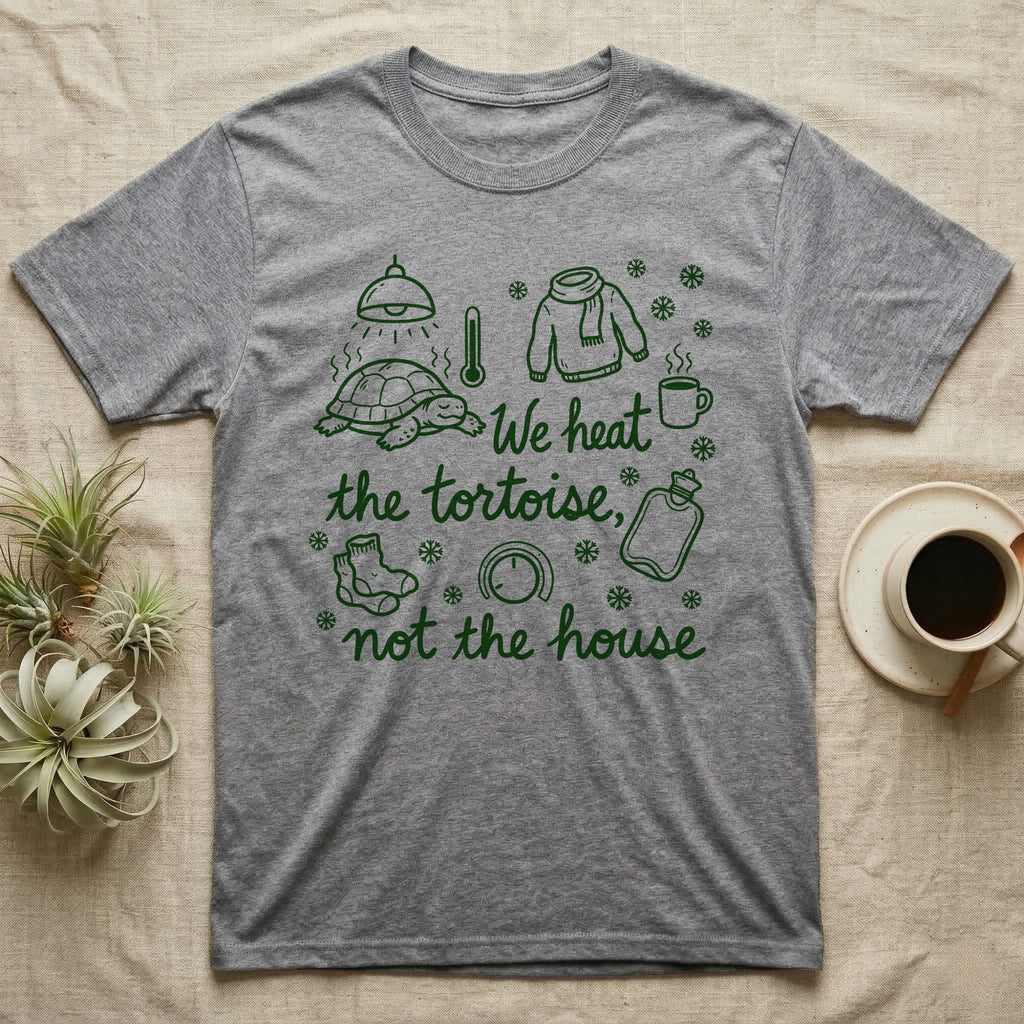 Russian Tortoise Winter T-Shirt in Sport Grey — Green marker cursive reading We heat the tortoise, not the house: a tortoise basking under a heat lamp while socks, scarves and snowflakes fill the cold house