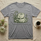 Grey t-shirt with frog illustration on rock with ferns and mushrooms.