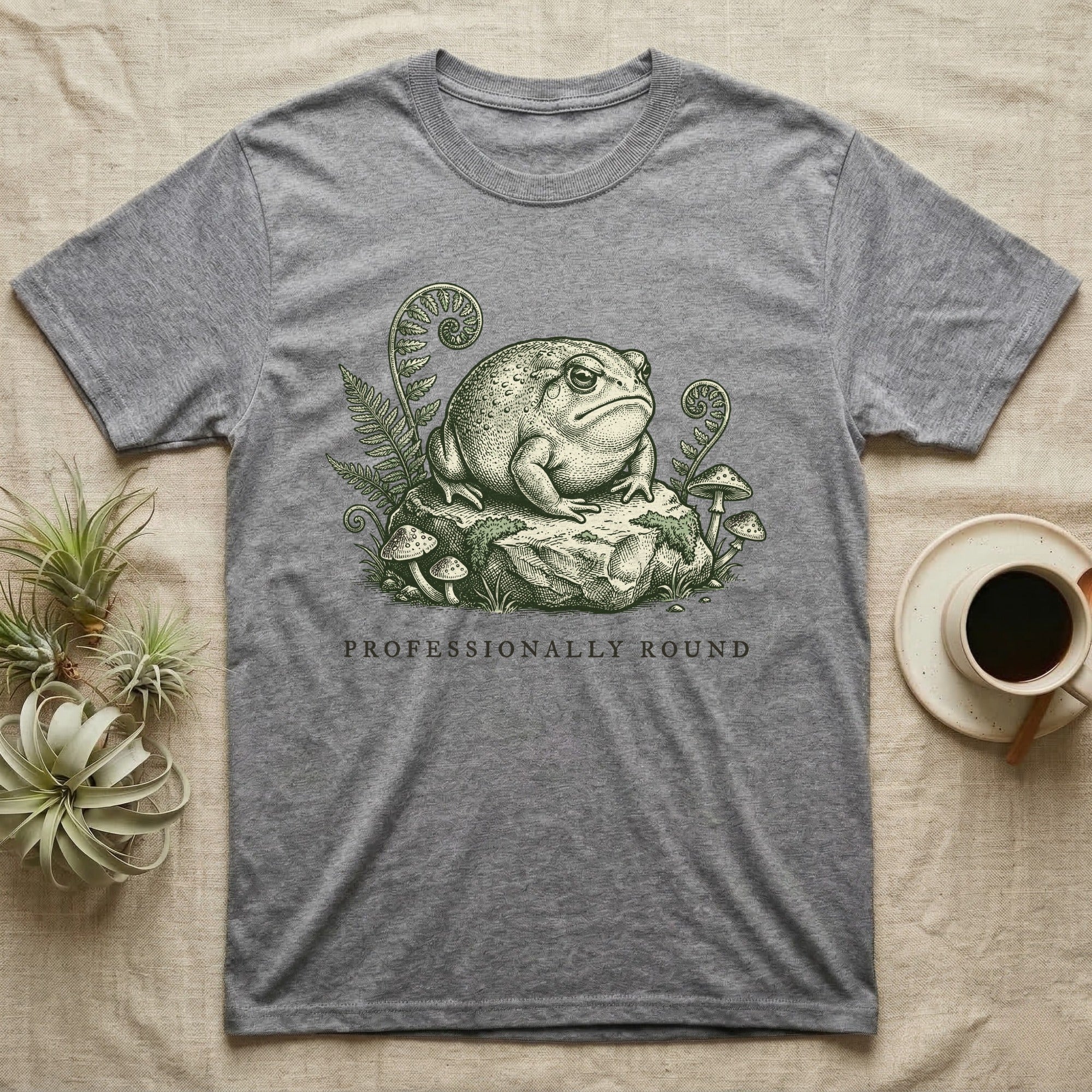 Grey t-shirt with frog illustration on rock with ferns and mushrooms.