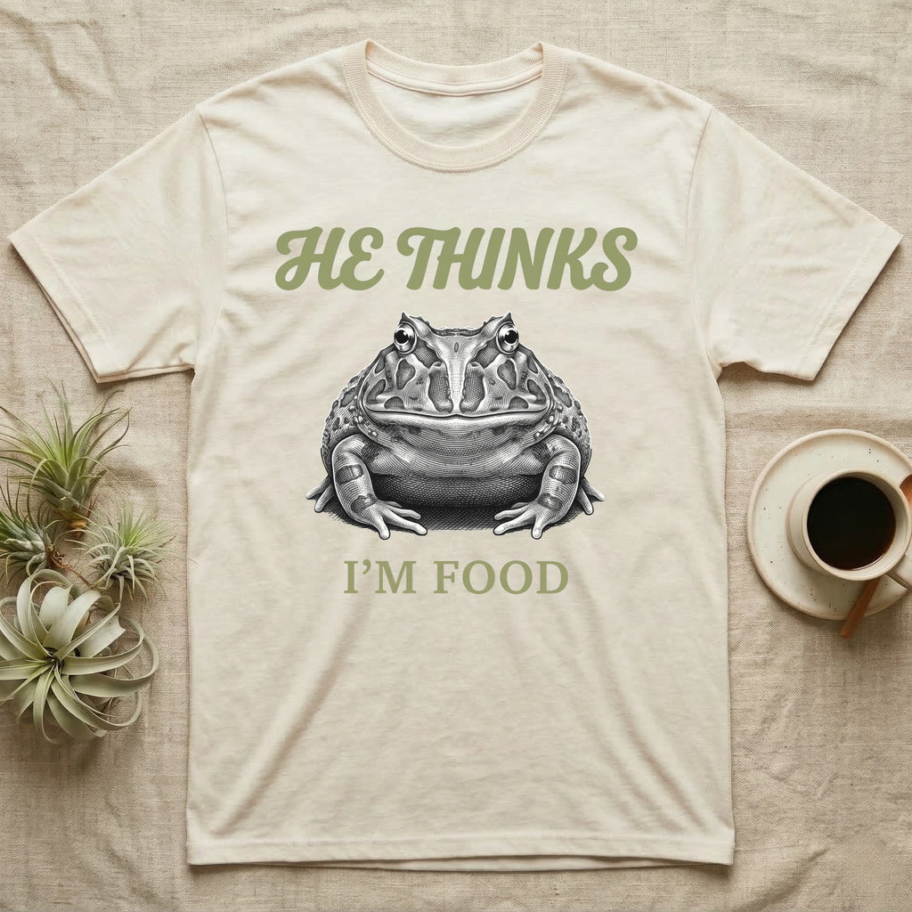 Pacman Frog He Thinks I'm Food T-Shirt in Natural — Engraved white-line illustration of a pacman frog staring straight ahead below retro script lettering reading HE THINKS.