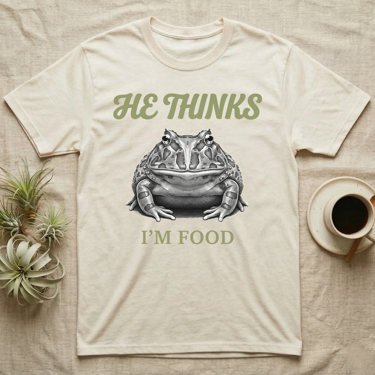 Pacman Frog He Thinks I'm Food T-Shirt in Natural — Engraved white-line illustration of a pacman frog staring straight ahead below retro script lettering reading HE THINKS.