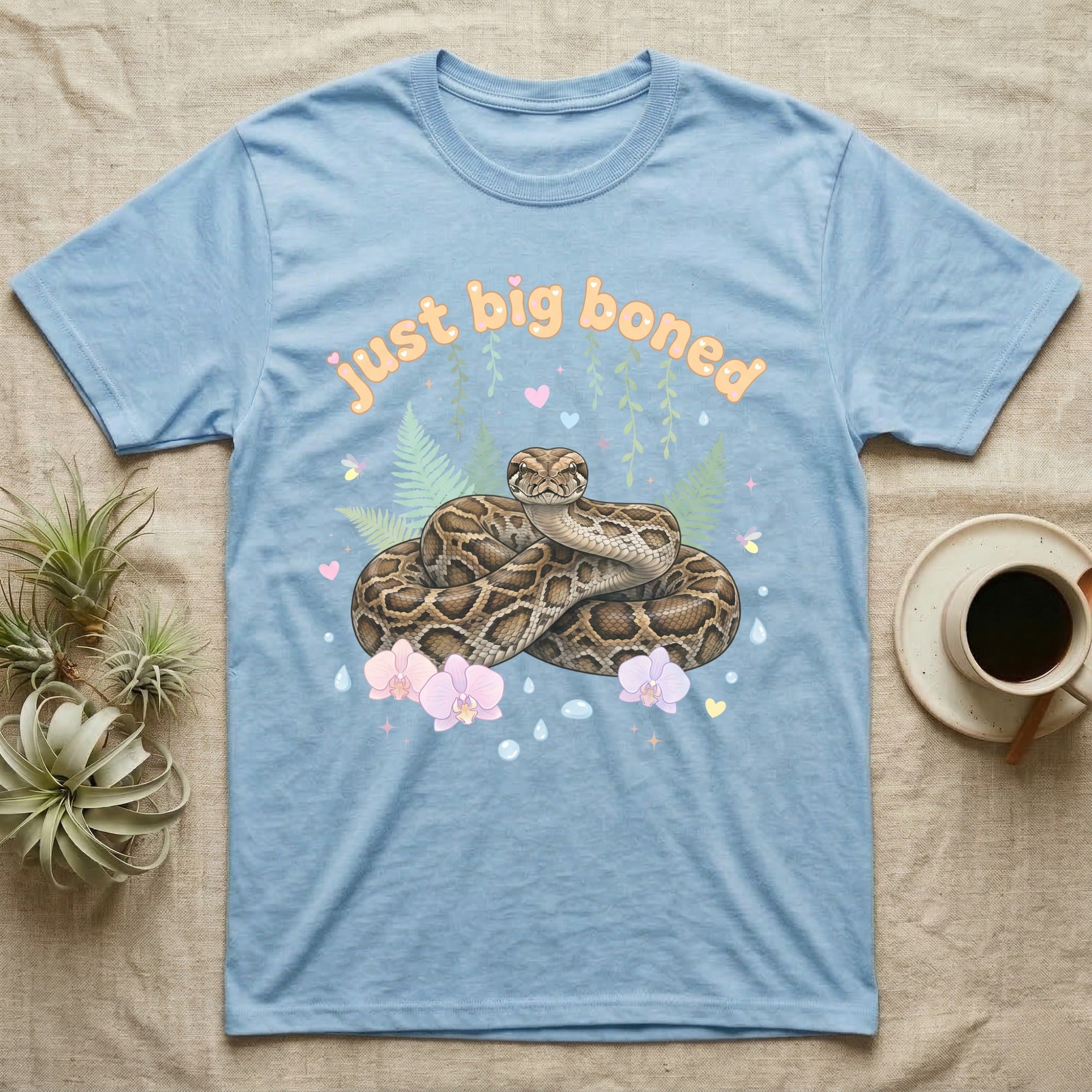 Burmese Python Baby T-Shirt in Light Blue — Massive coiled Burmese python among pastel ferns and orchids under puffy butterscotch bubble text "just big boned".