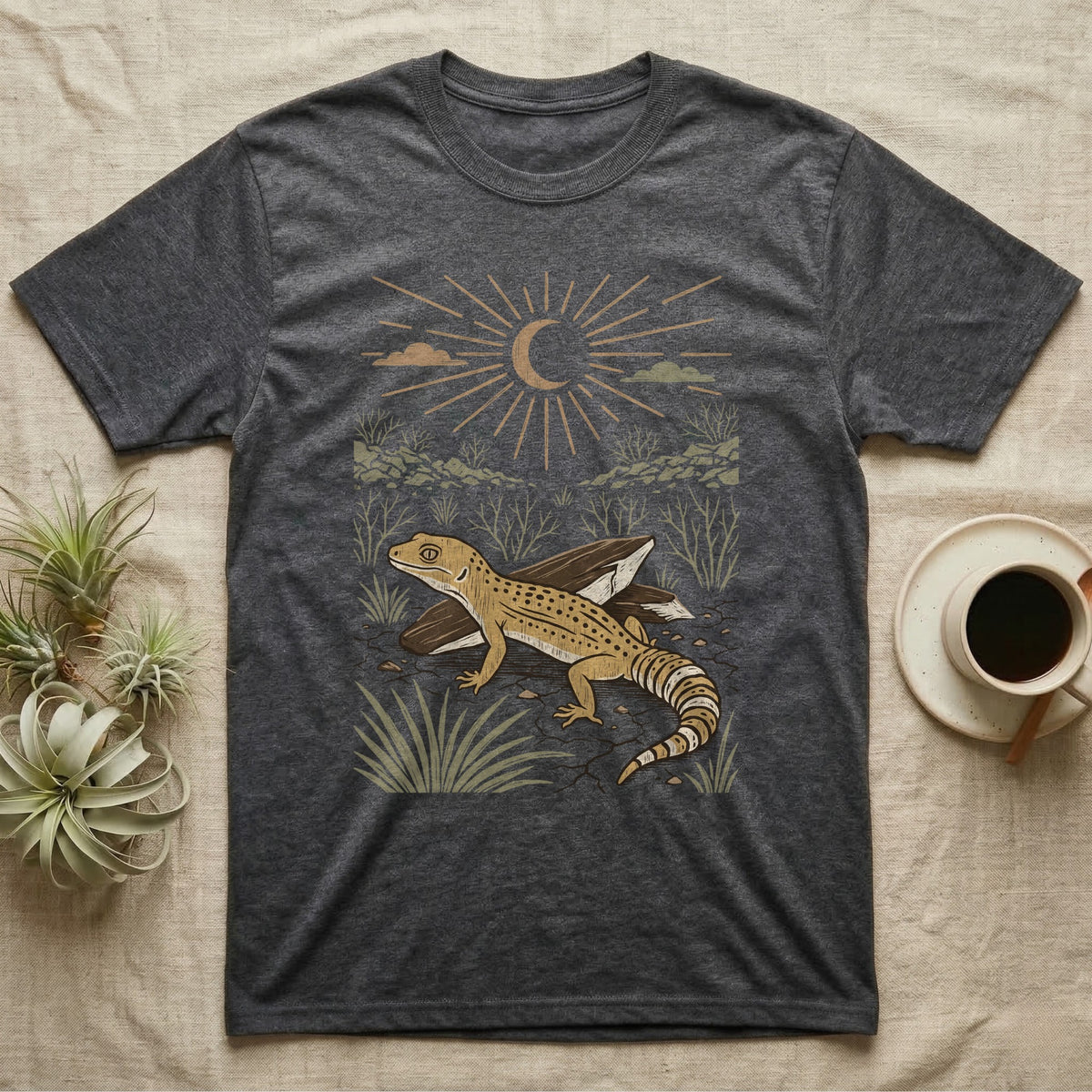 Leopard Gecko Home Range T-Shirt in Dark Heather — Woodcut-style illustration of a leopard gecko standing on cracked earth beside flat stones with thorn scrub behind, no on-shirt text.